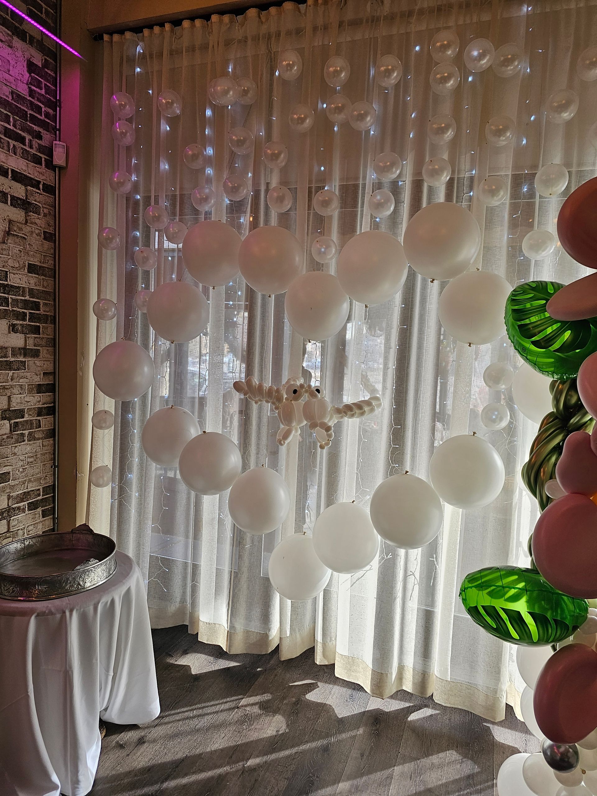 White balloons decorate a sheer curtain. Beside them are green and pink balloons, and a table with a white cloth.