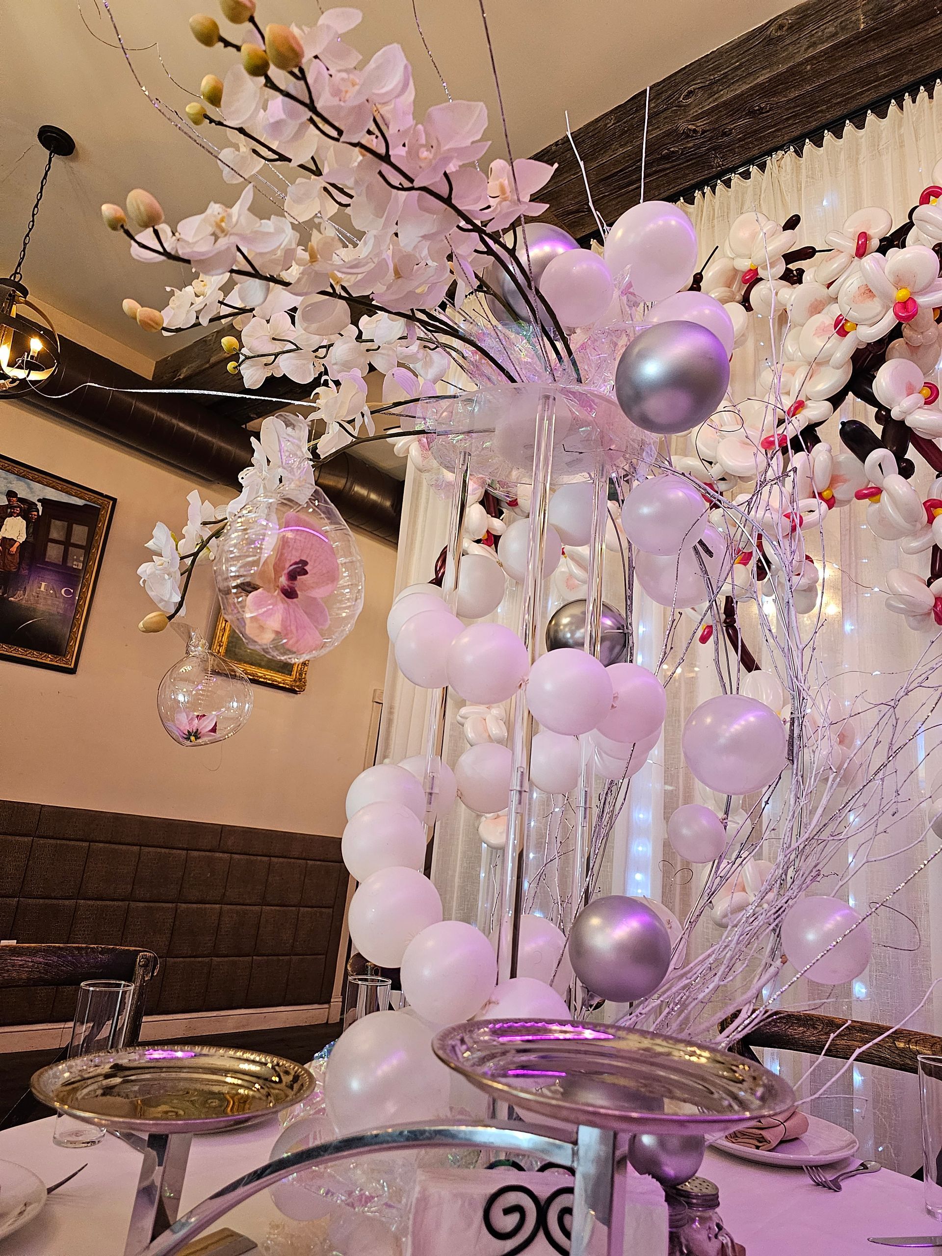 Elegant white and silver floral arrangement with balloons on a table, possibly for a celebration.
