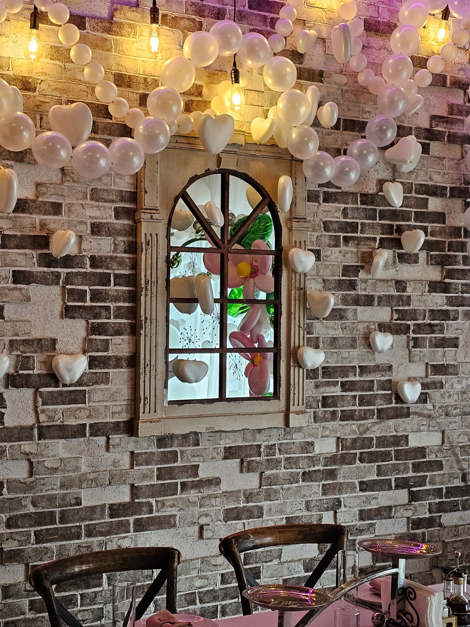 Brick wall with a window mirror. Decorated with white heart balloons, and string lights. Two chairs are in front.