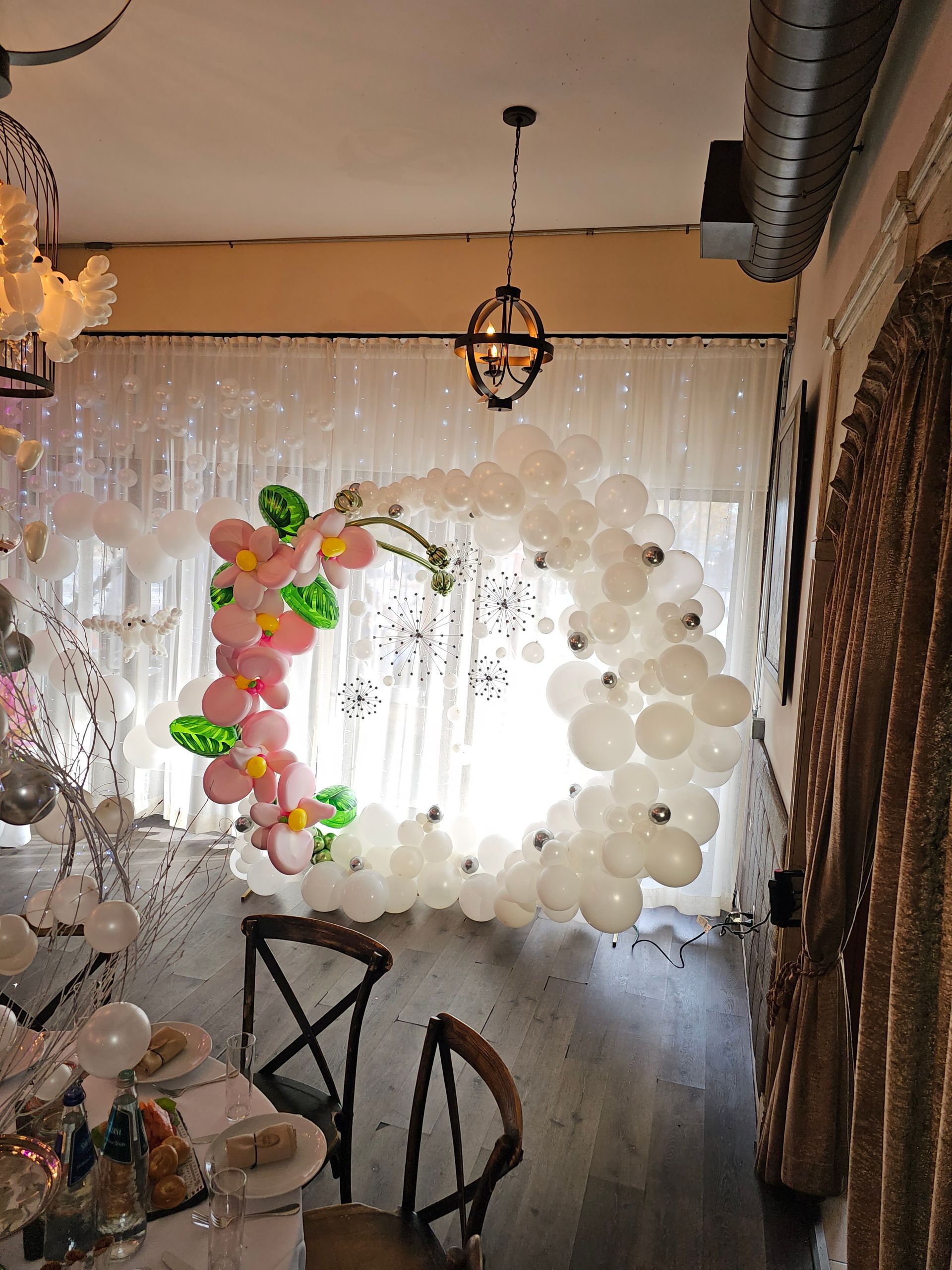 White balloon arch decorated with pink and green flowers inside a room with a table and chairs.