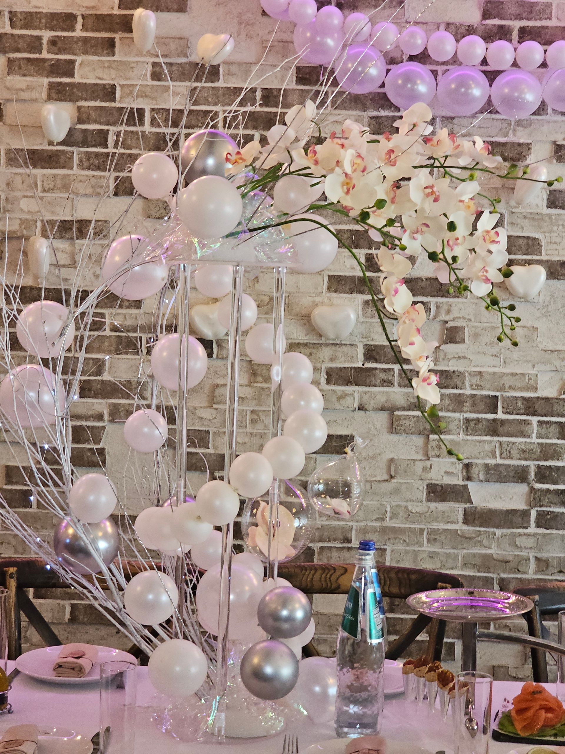 Elegant table setting with balloons, flowers, and silver accents against a brick wall background.