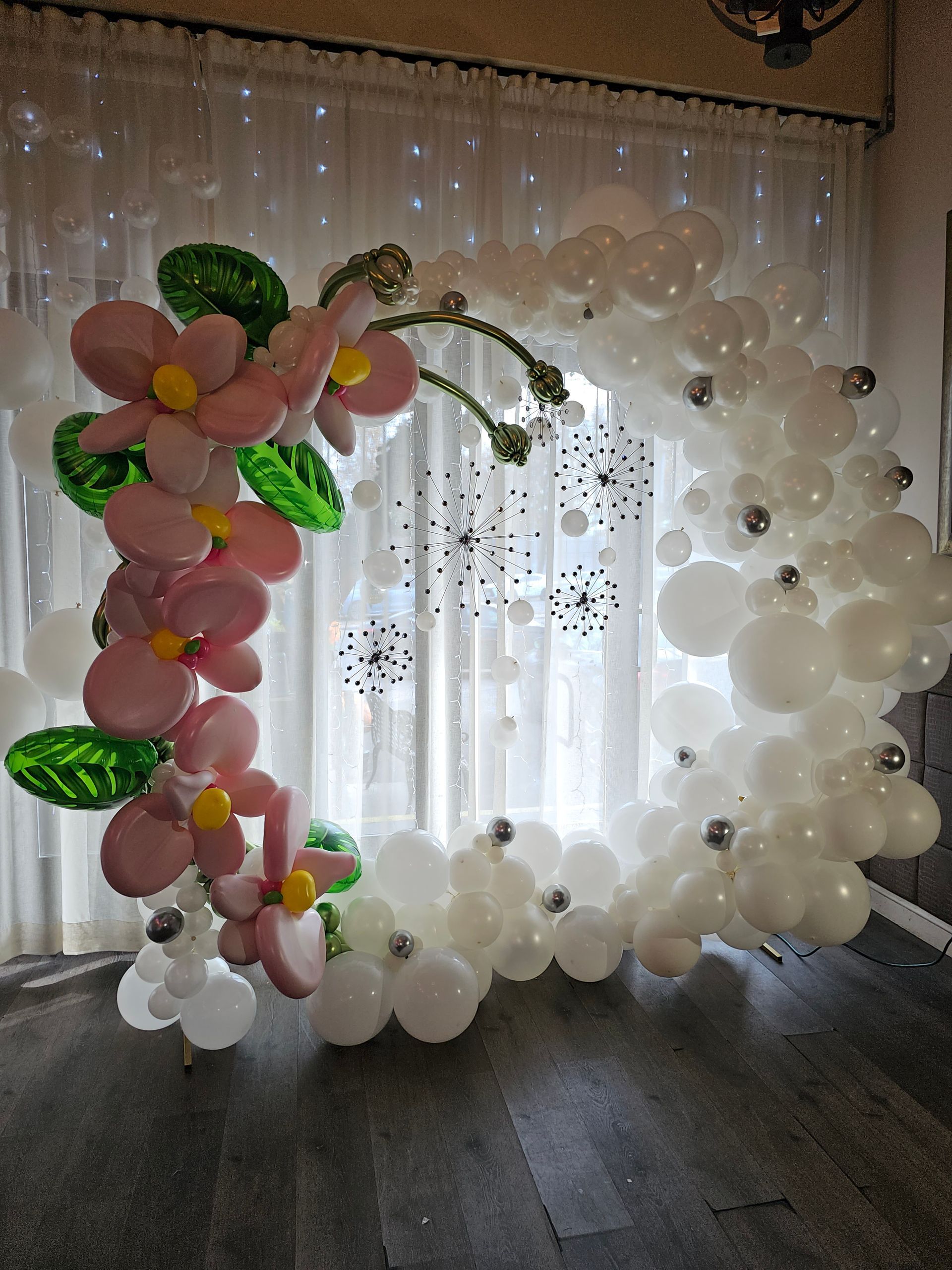 Balloon arch with pink flowers, green leaves, white and silver accents, set in front of a window.