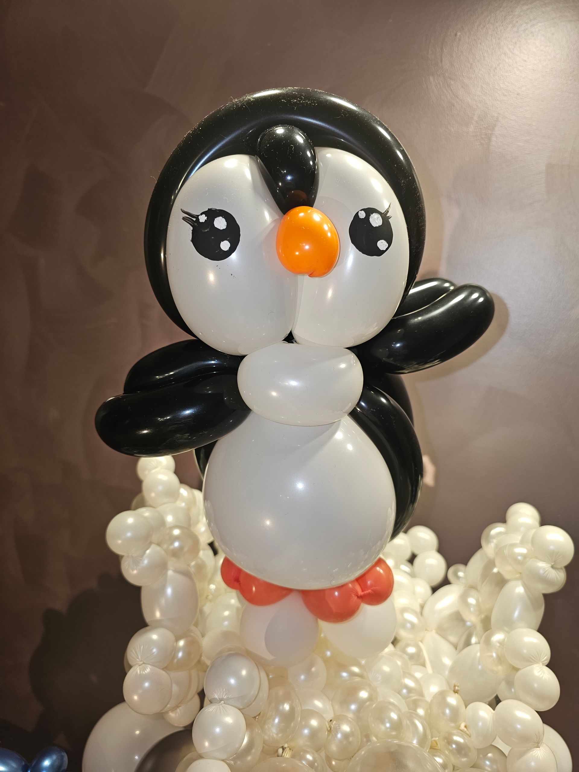 Balloon sculpture of a penguin with black and white body, orange beak, and large eyes, atop a snow-like base.