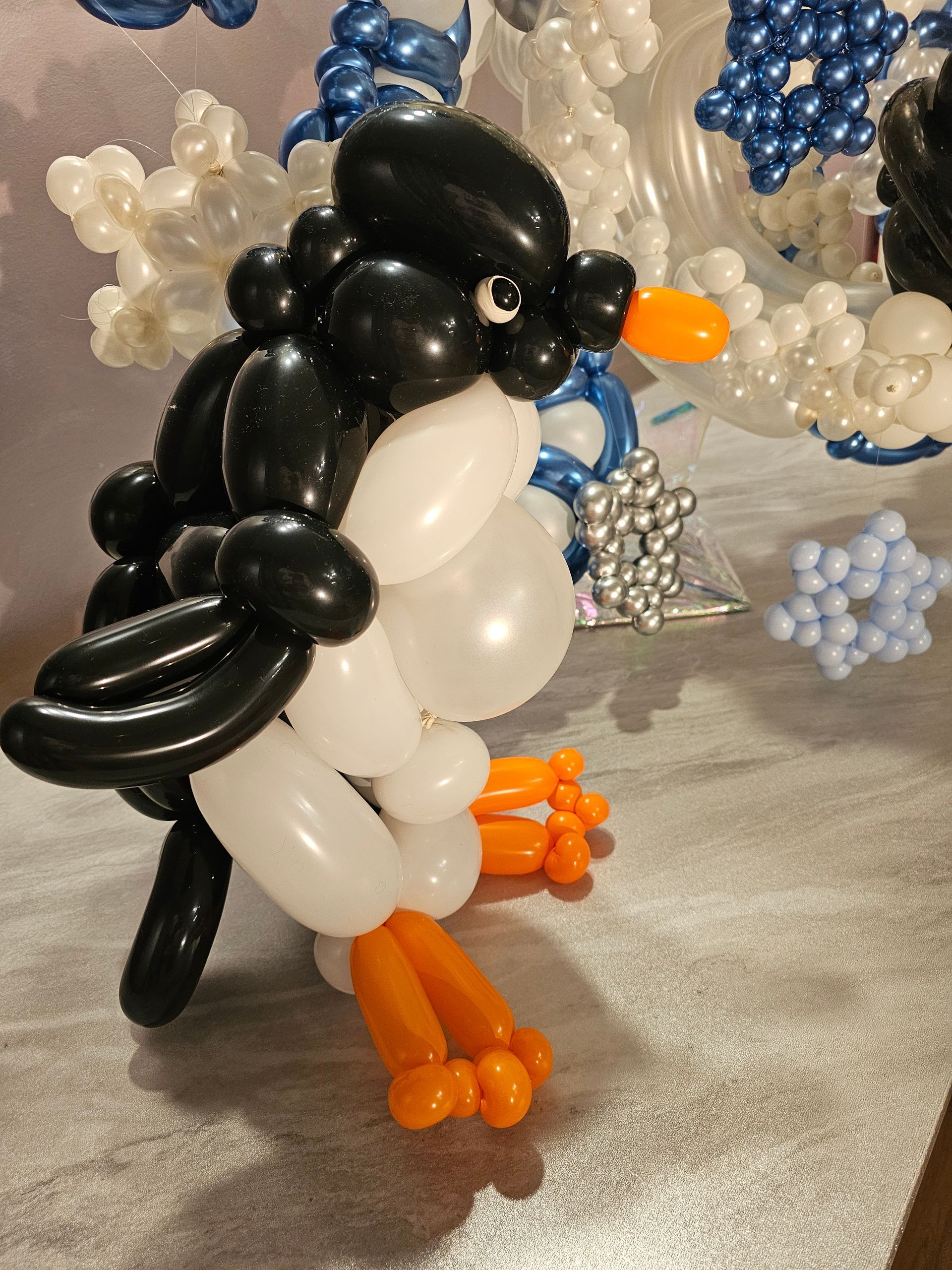 Balloon sculpture of a penguin with black and white body, orange beak and feet, set against a backdrop of snowflake balloons.