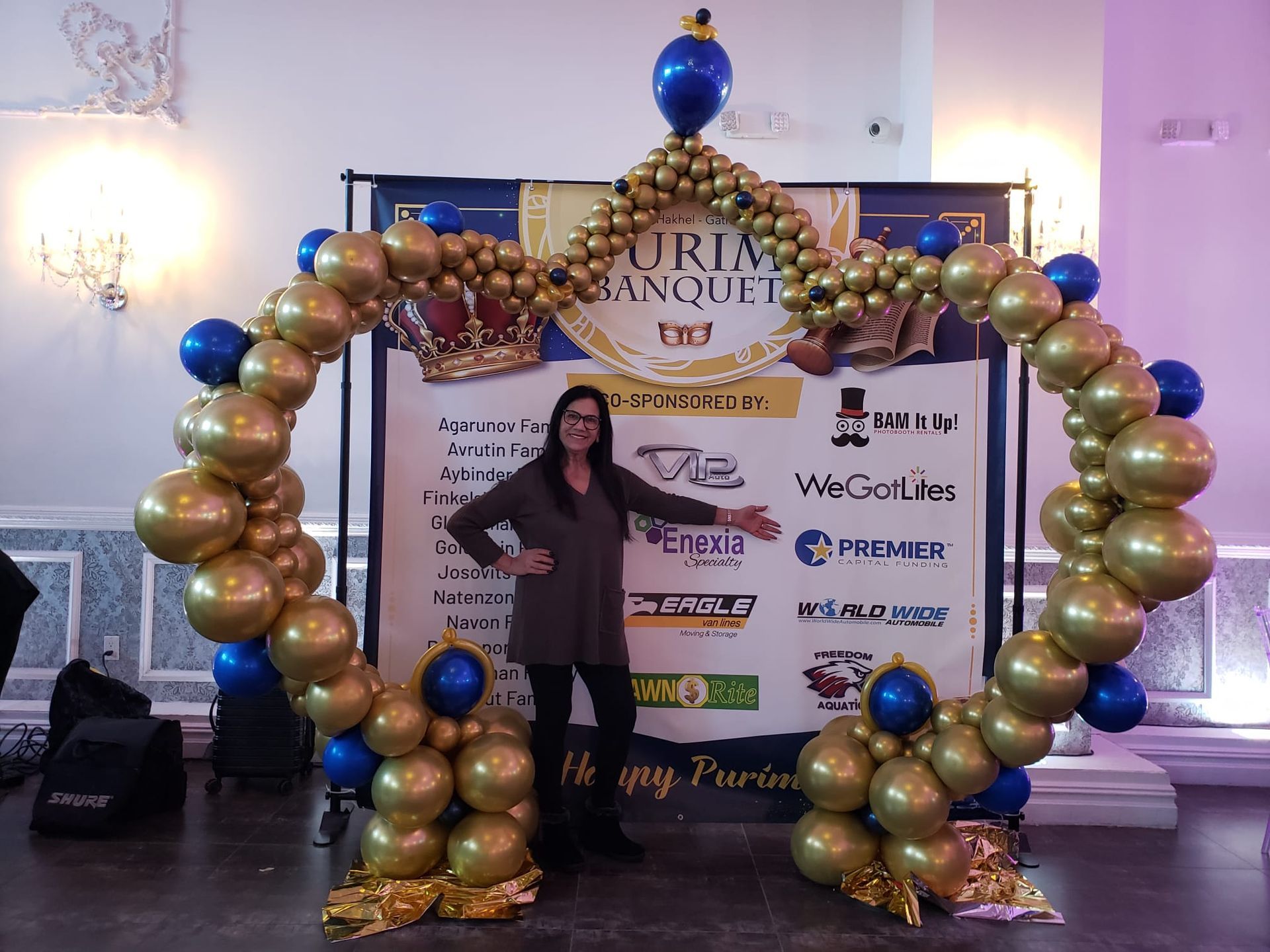 Woman stands under a gold and blue balloon arch at a Purim Banquet event.