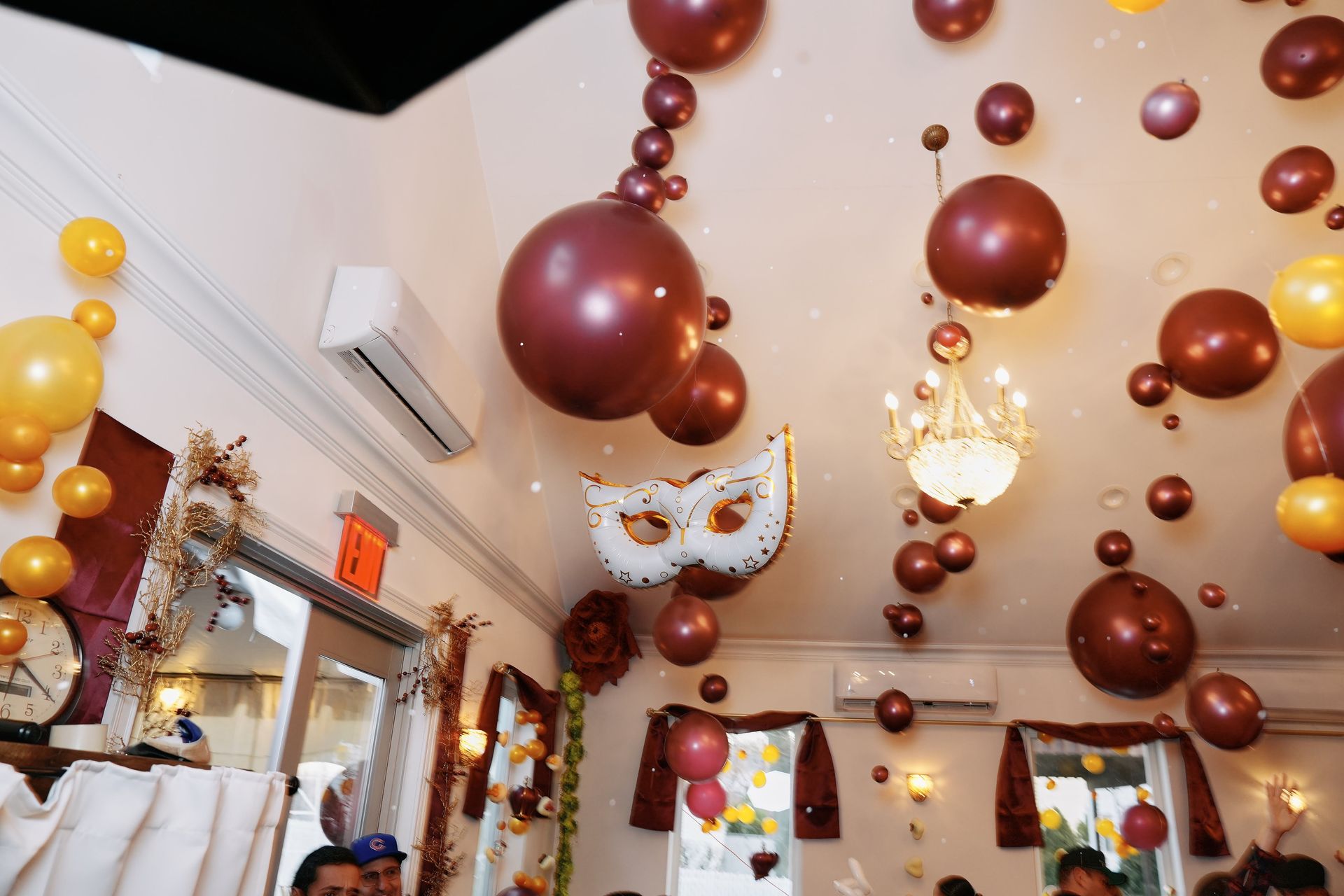 Room decorated with burgundy and gold balloons, a chandelier, and a mask for a celebration.