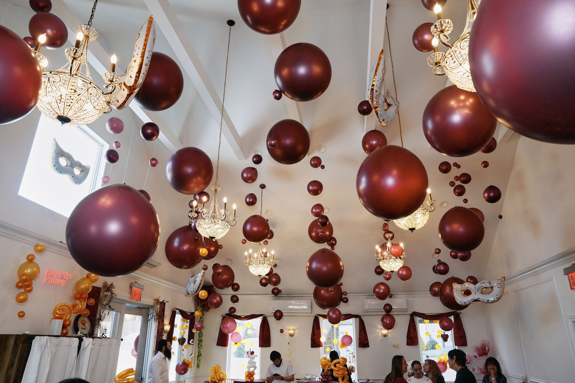 Burgundy balloons and chandeliers decorate a party venue ceiling.