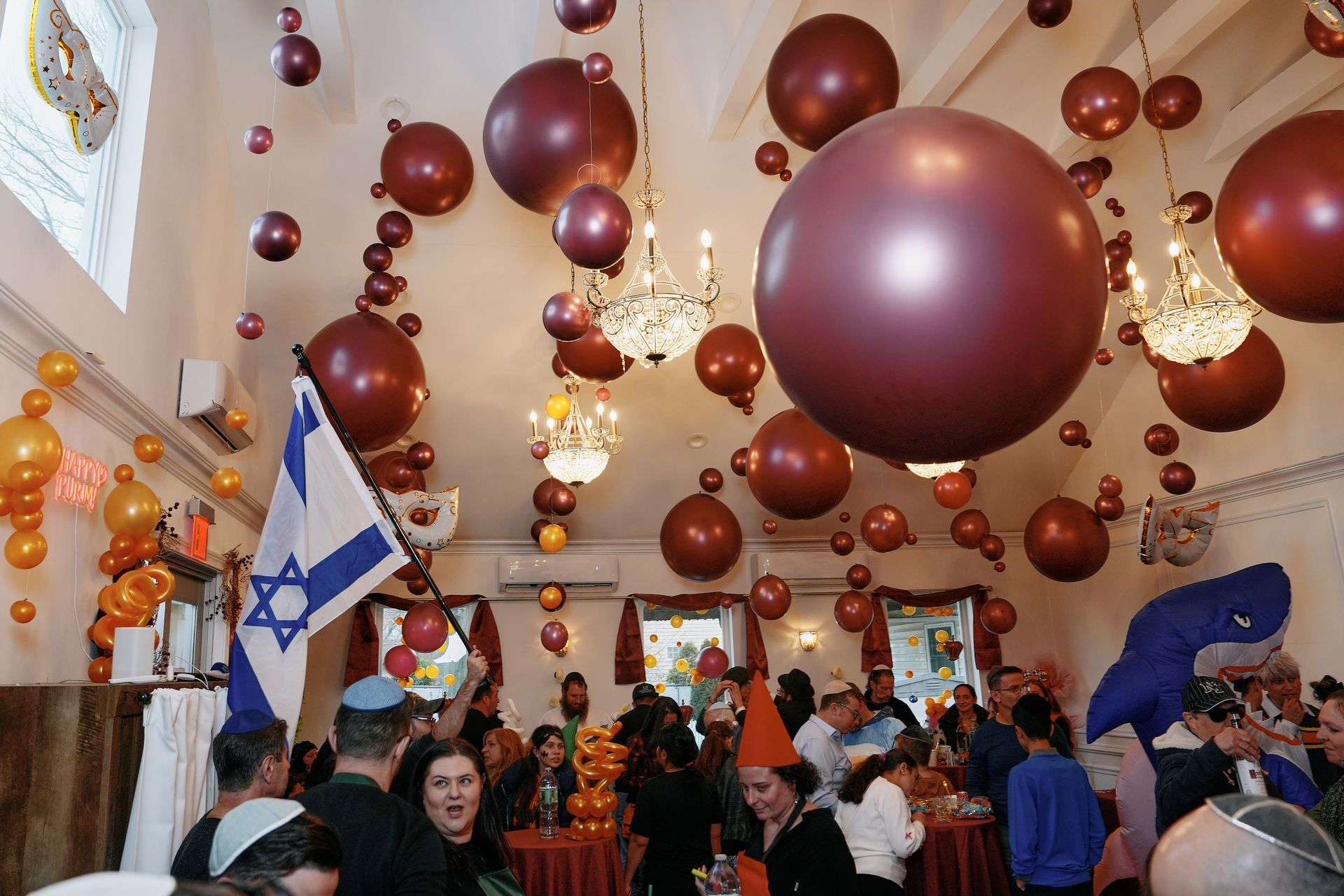 Party scene with burgundy balloons, Israeli flag, people gathered, chandeliers.