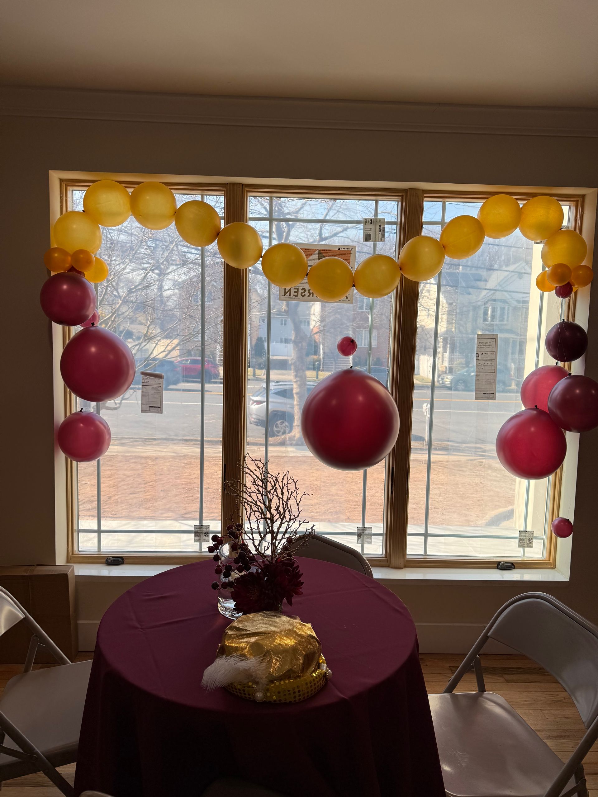 Table with cake and balloons, decorated in yellow and pink, in front of a window.