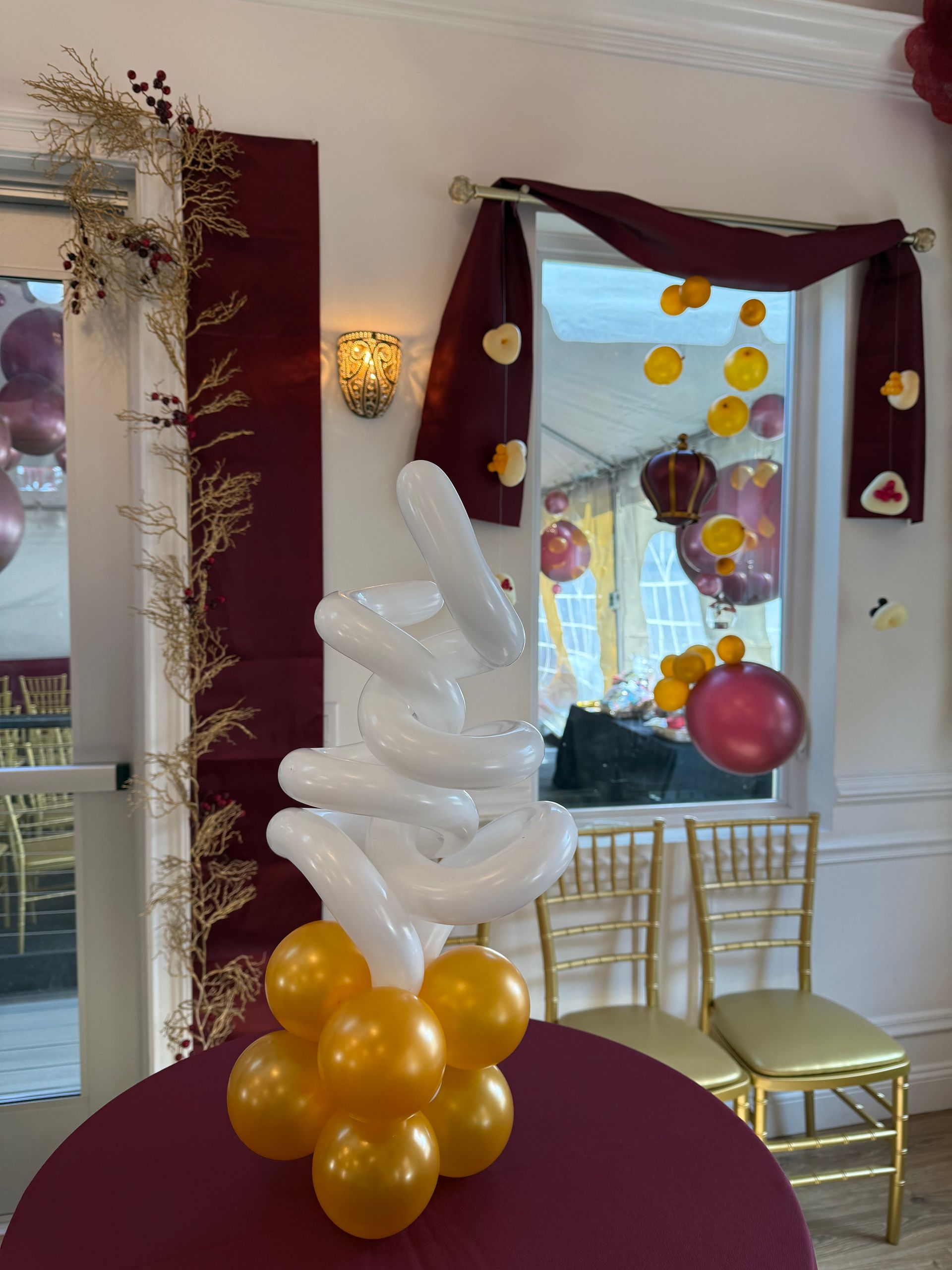 Balloon decorations at an event: gold and white balloons on a table with a maroon tablecloth, window in background.