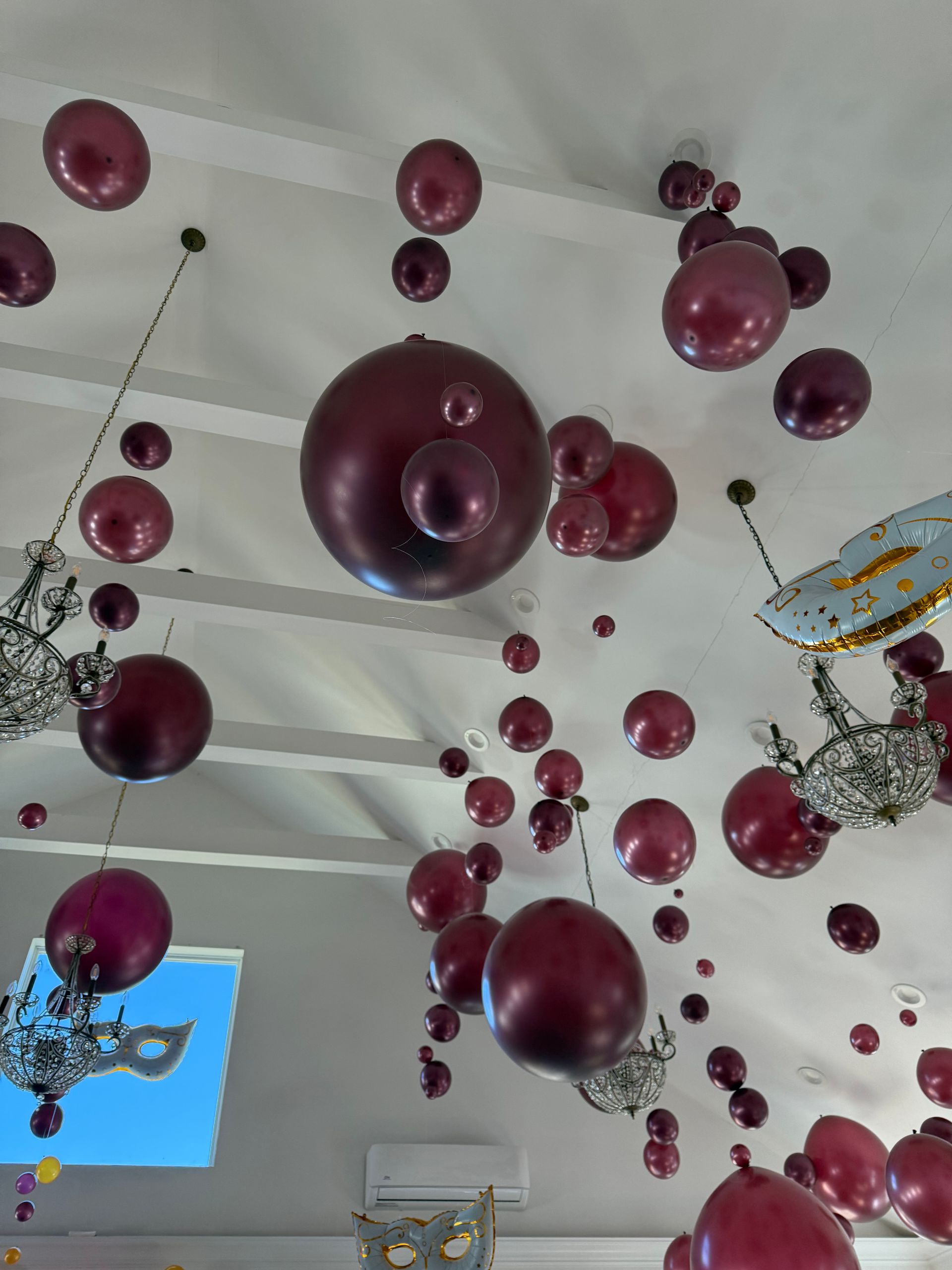 Maroon balloons of varying sizes suspended from a white ceiling, alongside decorative chandeliers.