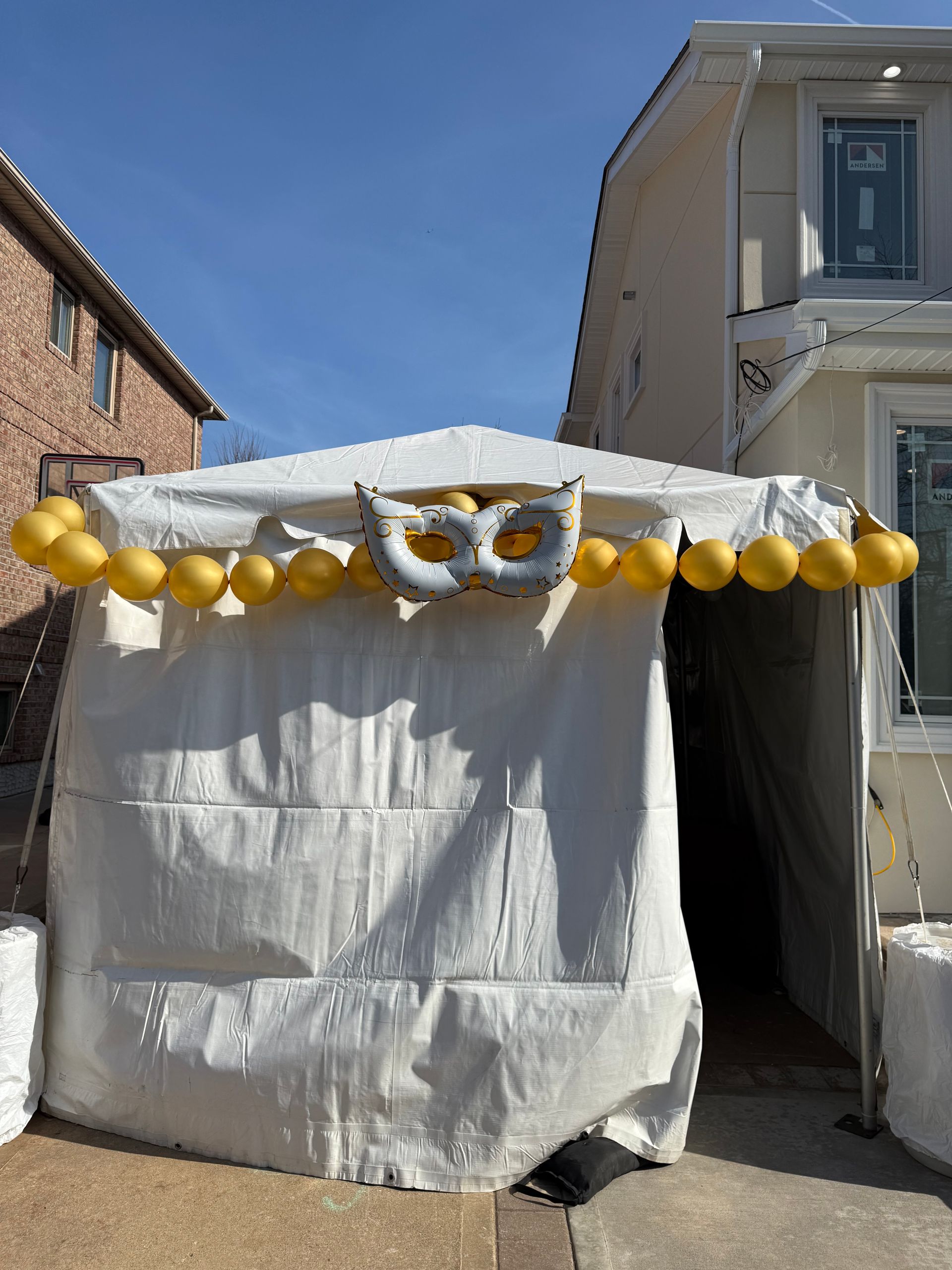White tent with gold balloons and a silver mask decoration, set outdoors.