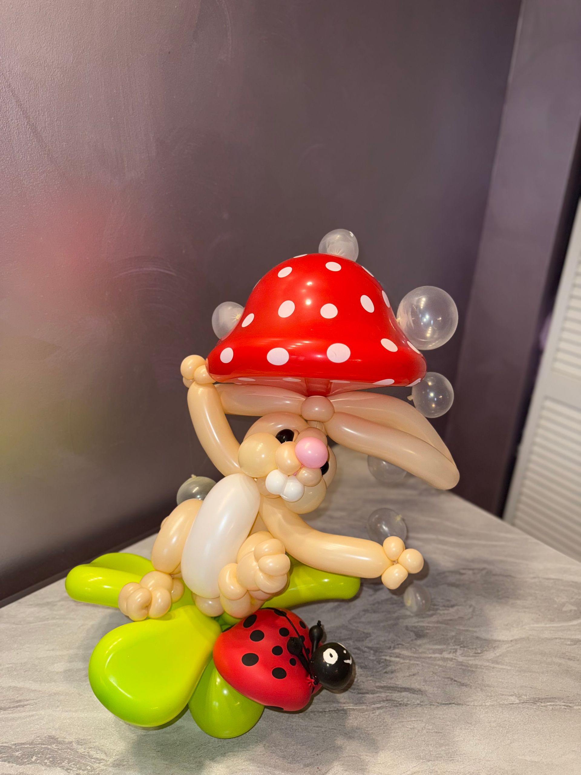 Balloon sculpture of a rabbit under a red mushroom, with a ladybug on a green flower.