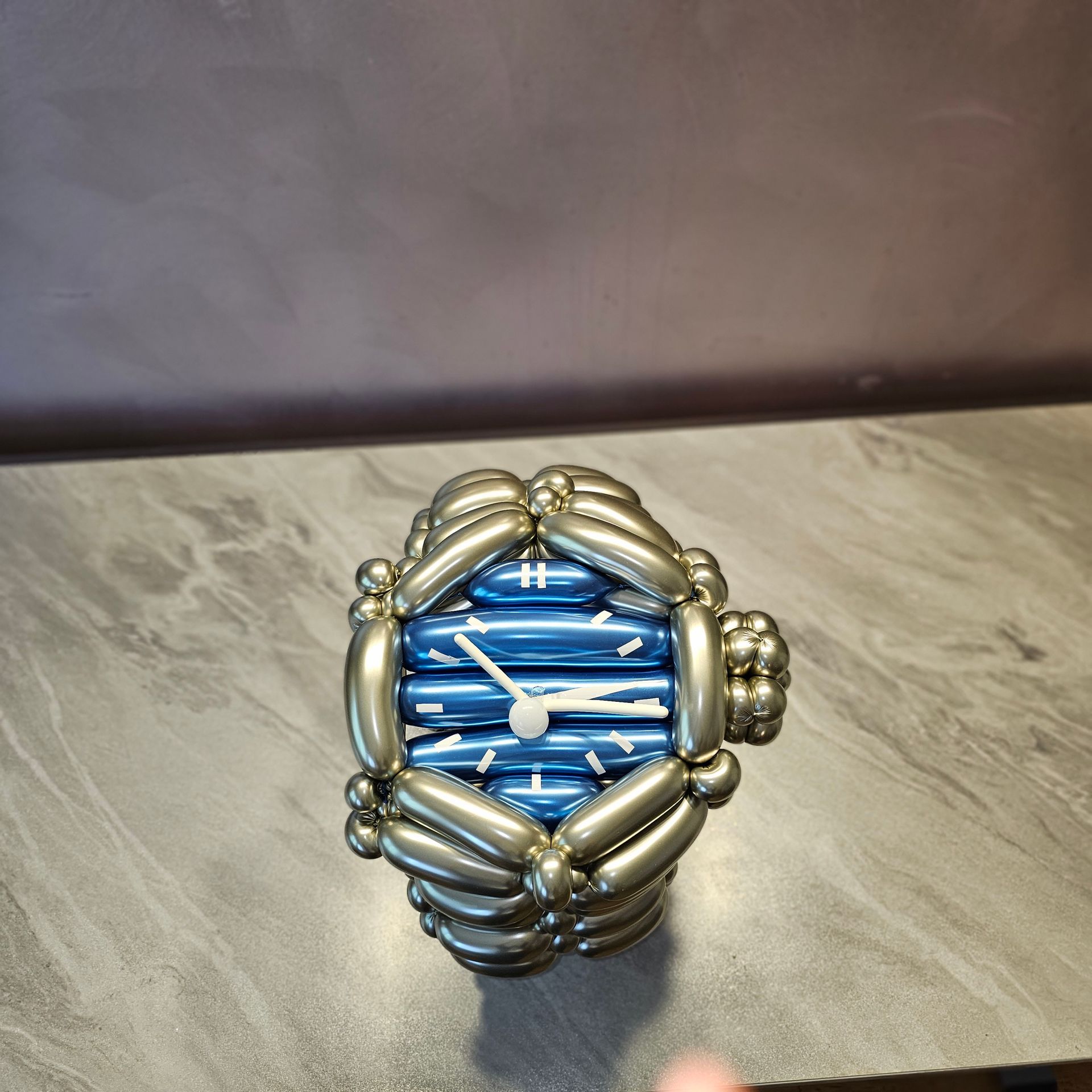 A watch made of silver balloons with a blue watch face on a gray surface.