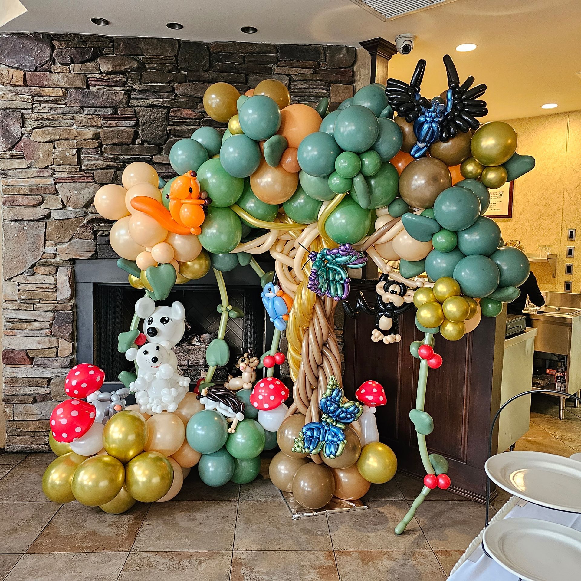 Balloon tree sculpture with woodland animals and colorful balloons, in front of a fireplace.