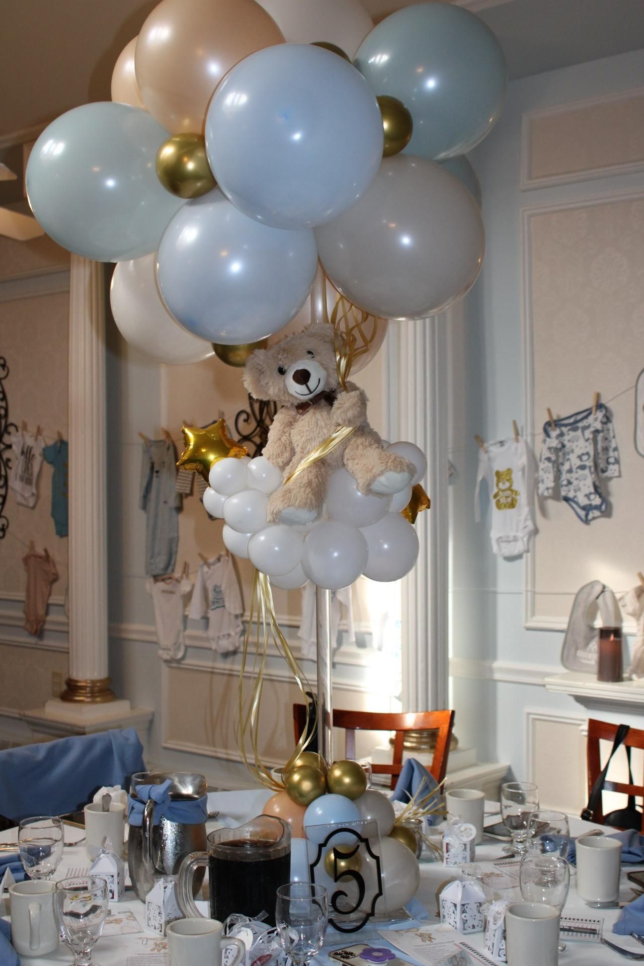 Balloon centerpiece with teddy bear, clouds, and gold accents on a table set for a baby shower.