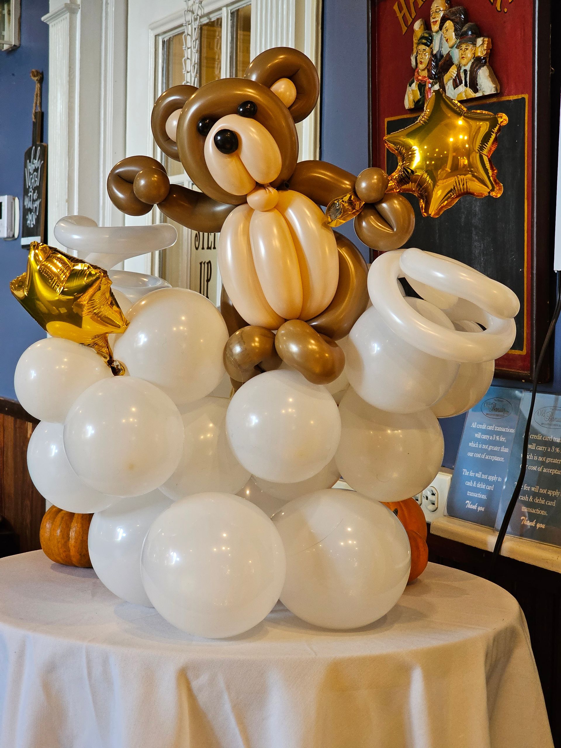 Balloon sculpture of a brown bear, white balloons, and gold stars on a table.