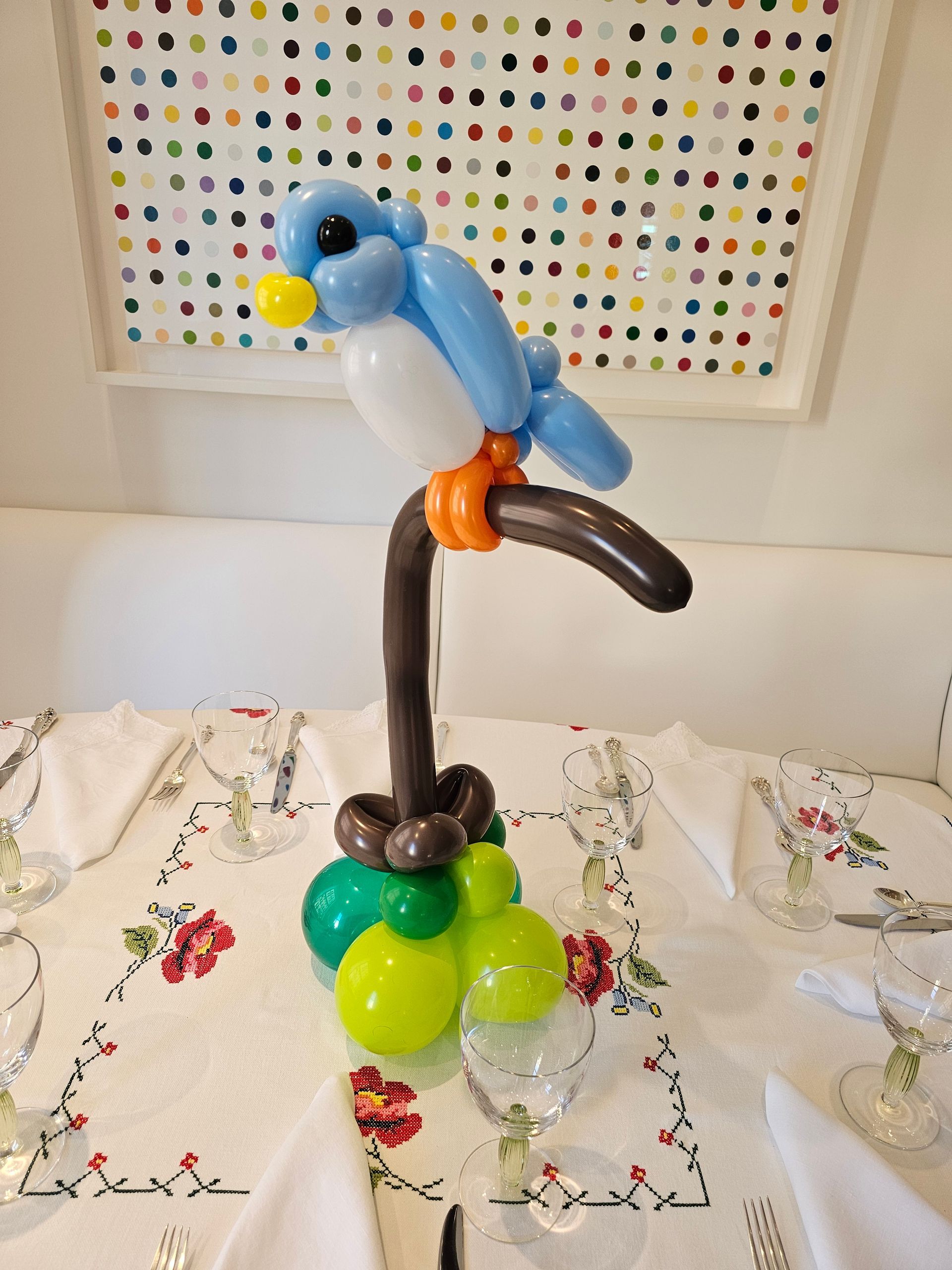 Blue balloon bird perched on a brown balloon branch, over a green and brown base. Table setting.