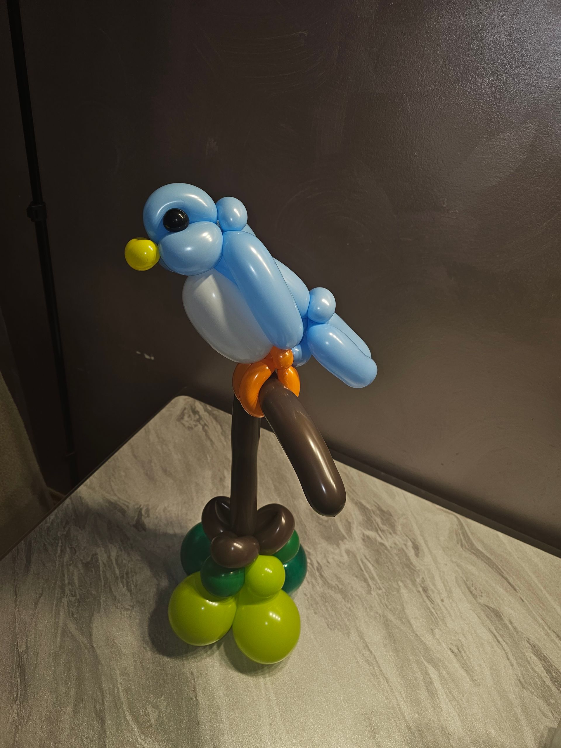 Balloon sculpture of a blue bird perched on a brown branch with a green and brown base.