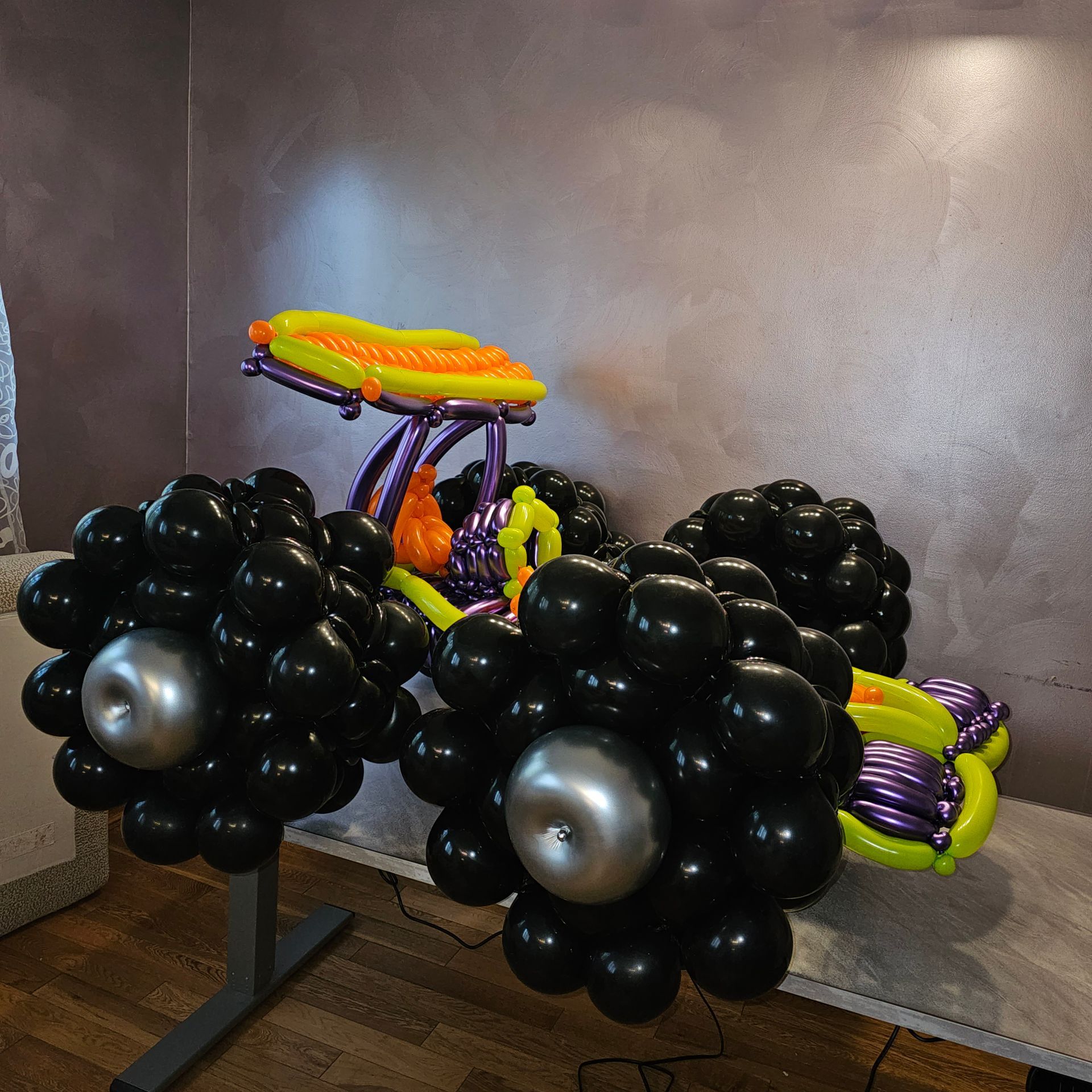 Balloon sculpture of a black car with silver wheel centers, and a yellow, orange, and purple superstructure.