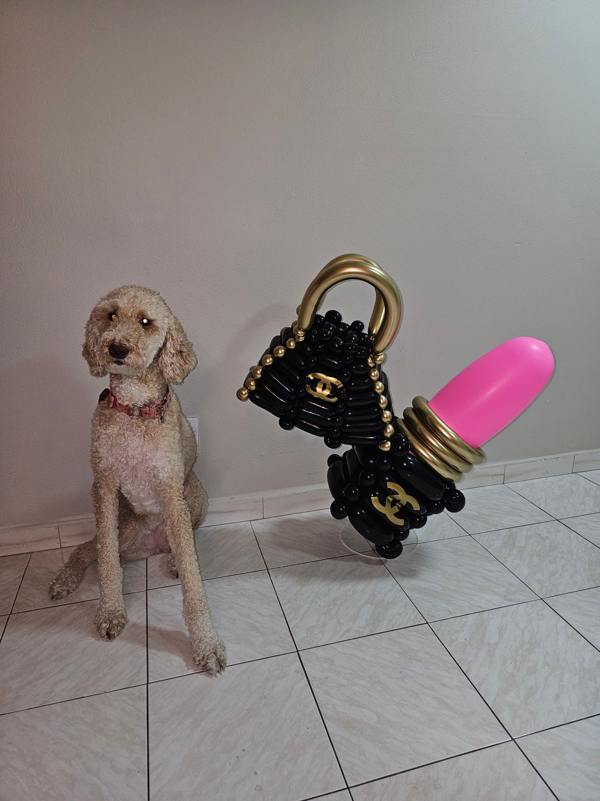 Dog sits near a black purse charm with lipstick on a tiled floor.