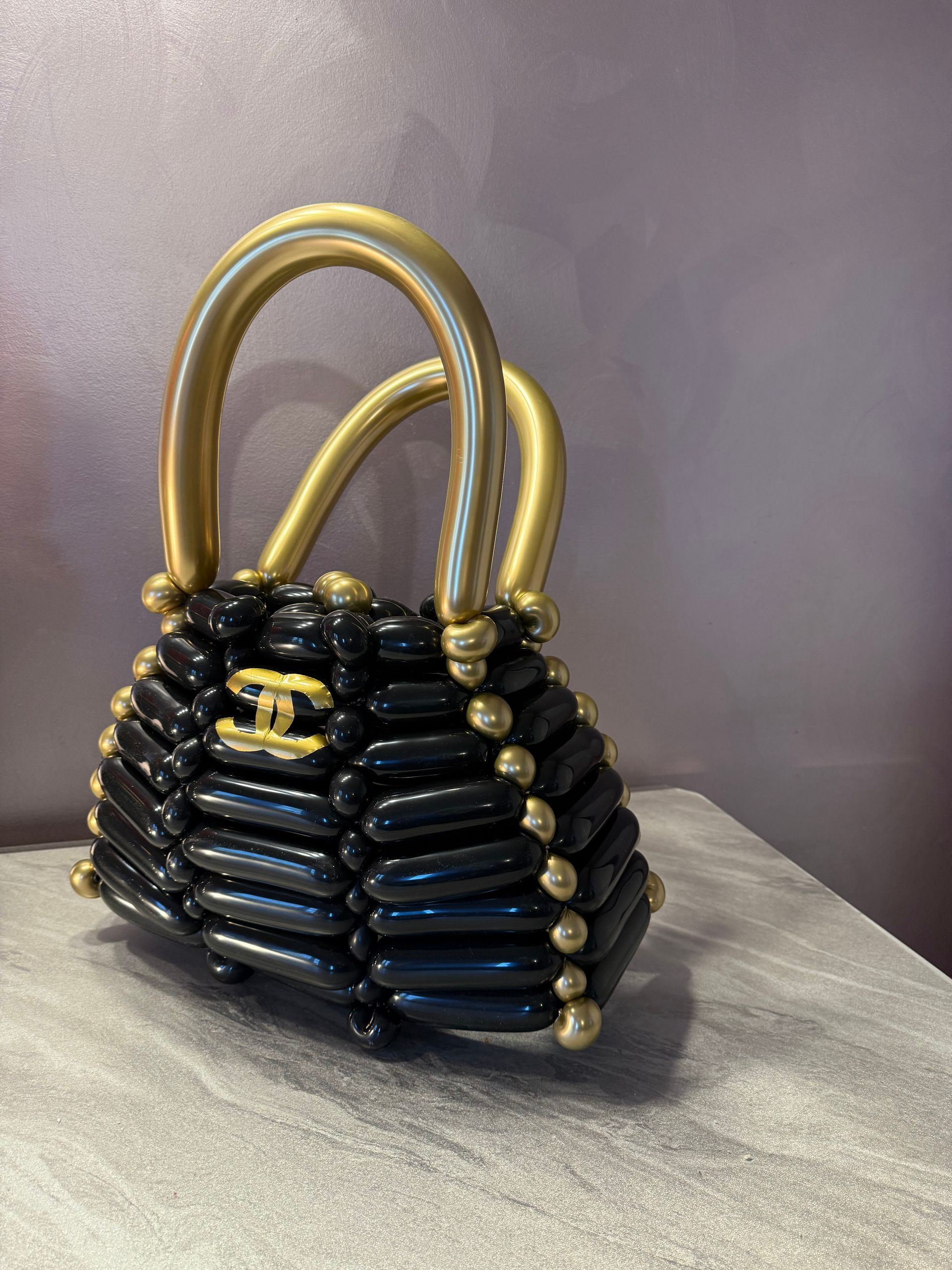 Black and gold balloon sculpture of a handbag, with a gold logo and handles.