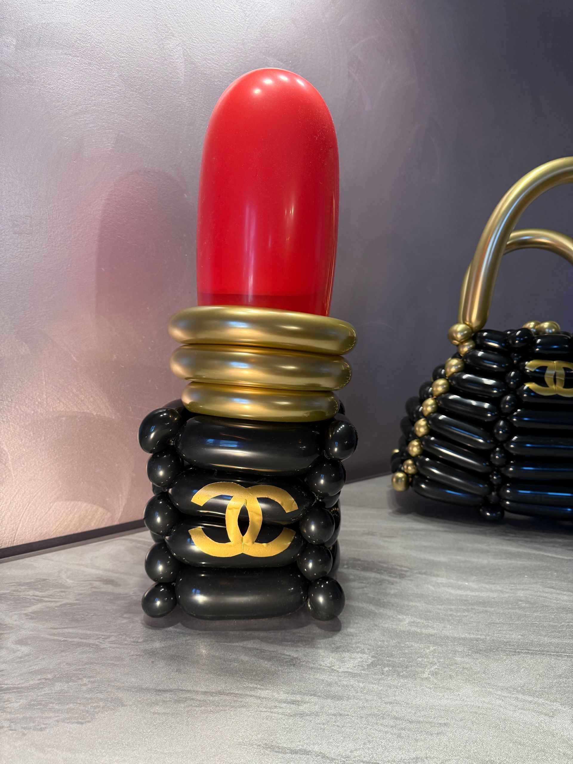 Balloon sculpture of a red lipstick with gold and black accents, next to a black handbag sculpture.
