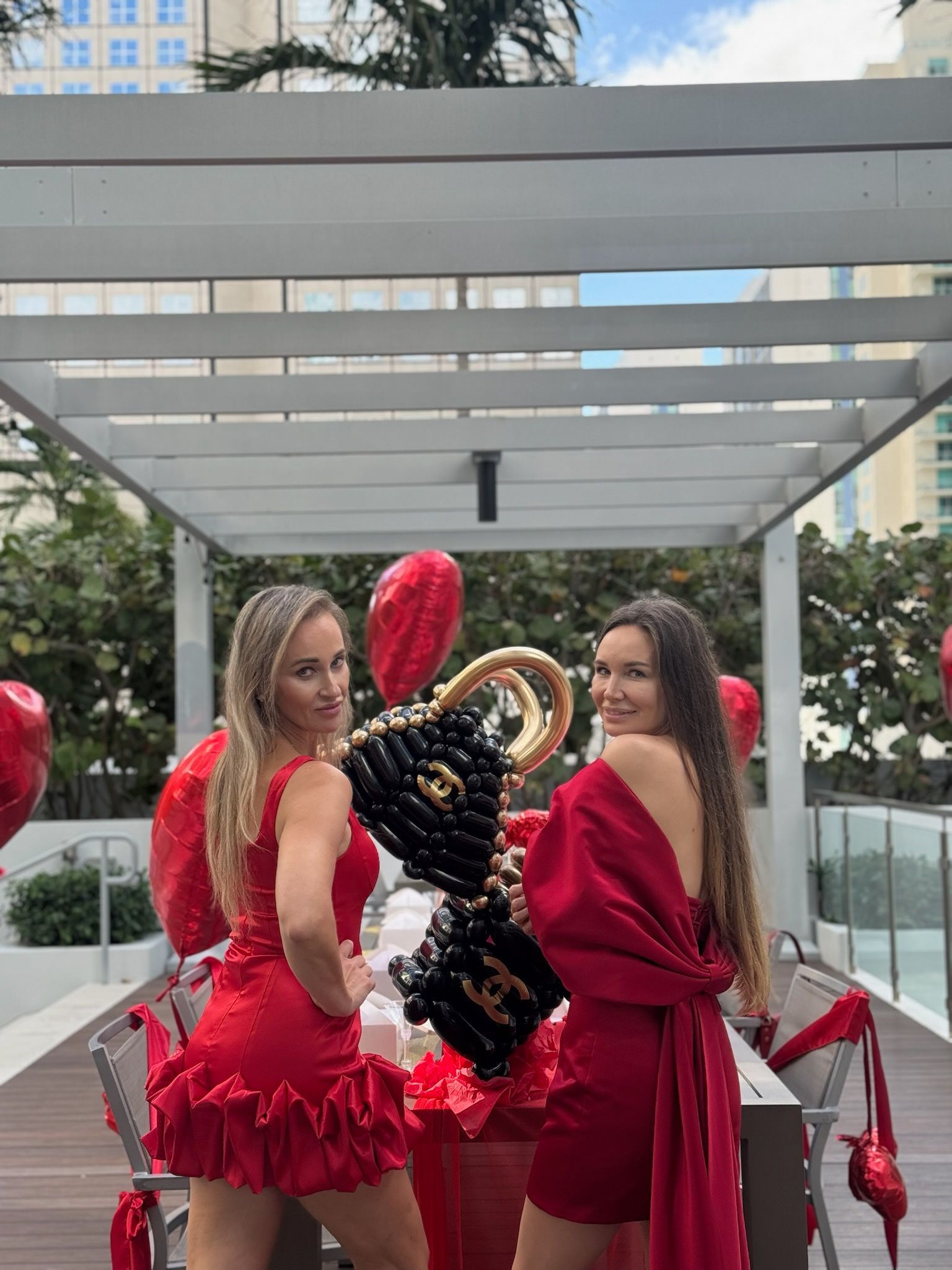 Two women in red dresses pose with heart-shaped balloons outdoors.