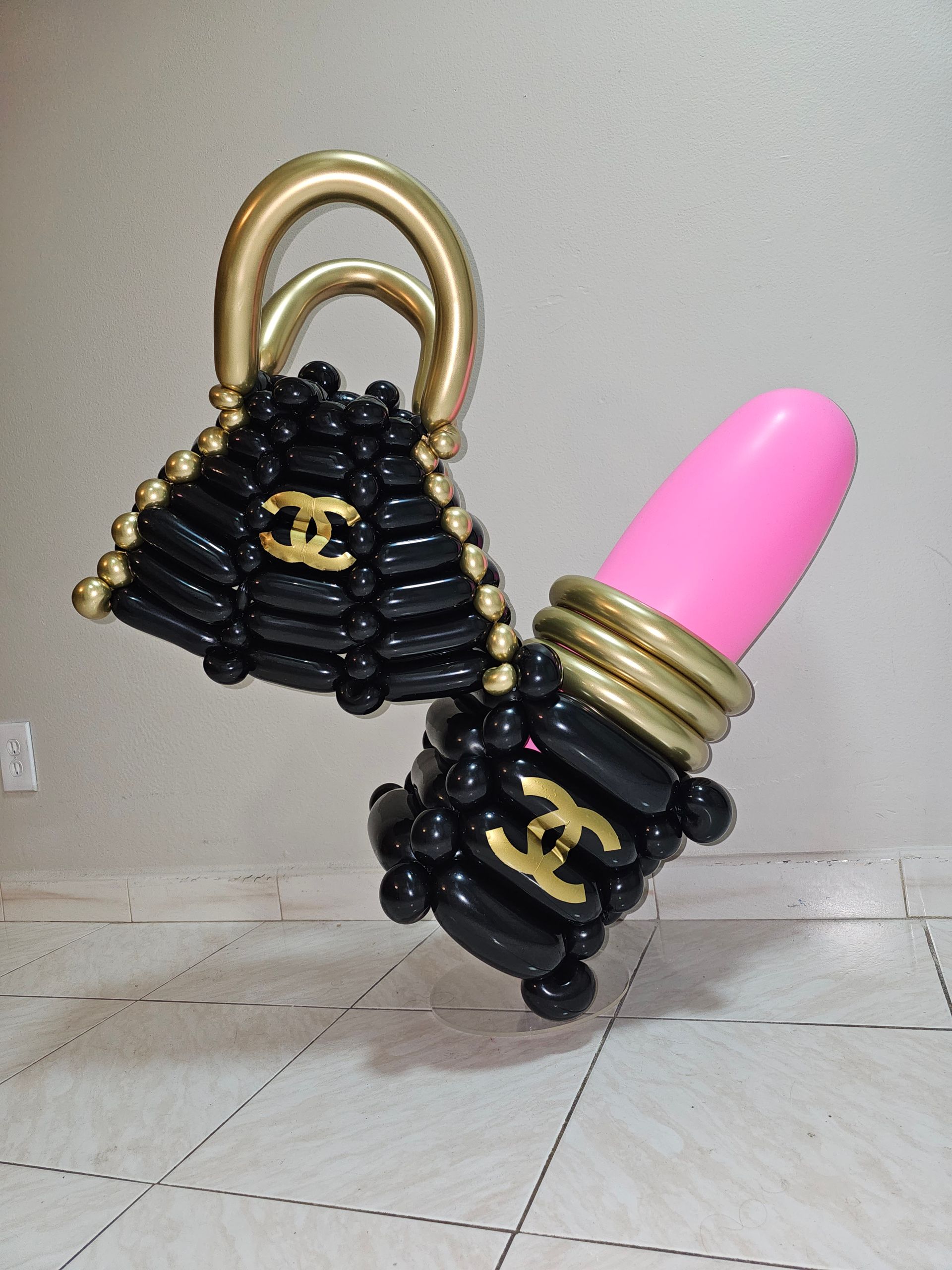 Black balloon purse and lipstick sculptures with gold accents and pink lipstick, on a tiled floor.