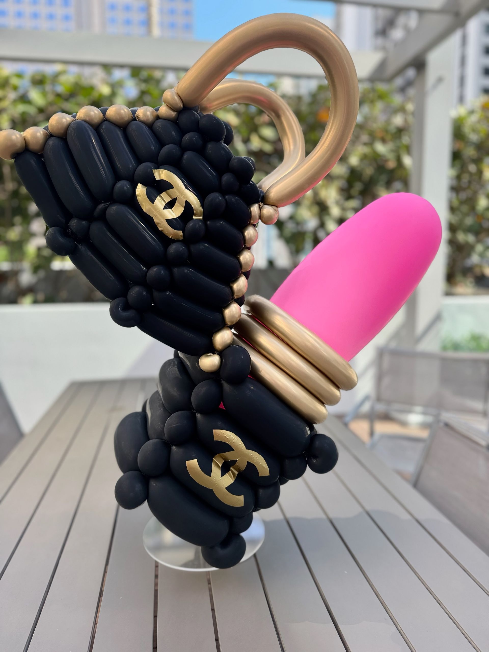 Balloon sculpture of a black handbag with a pink lipstick and Chanel logo on a table.