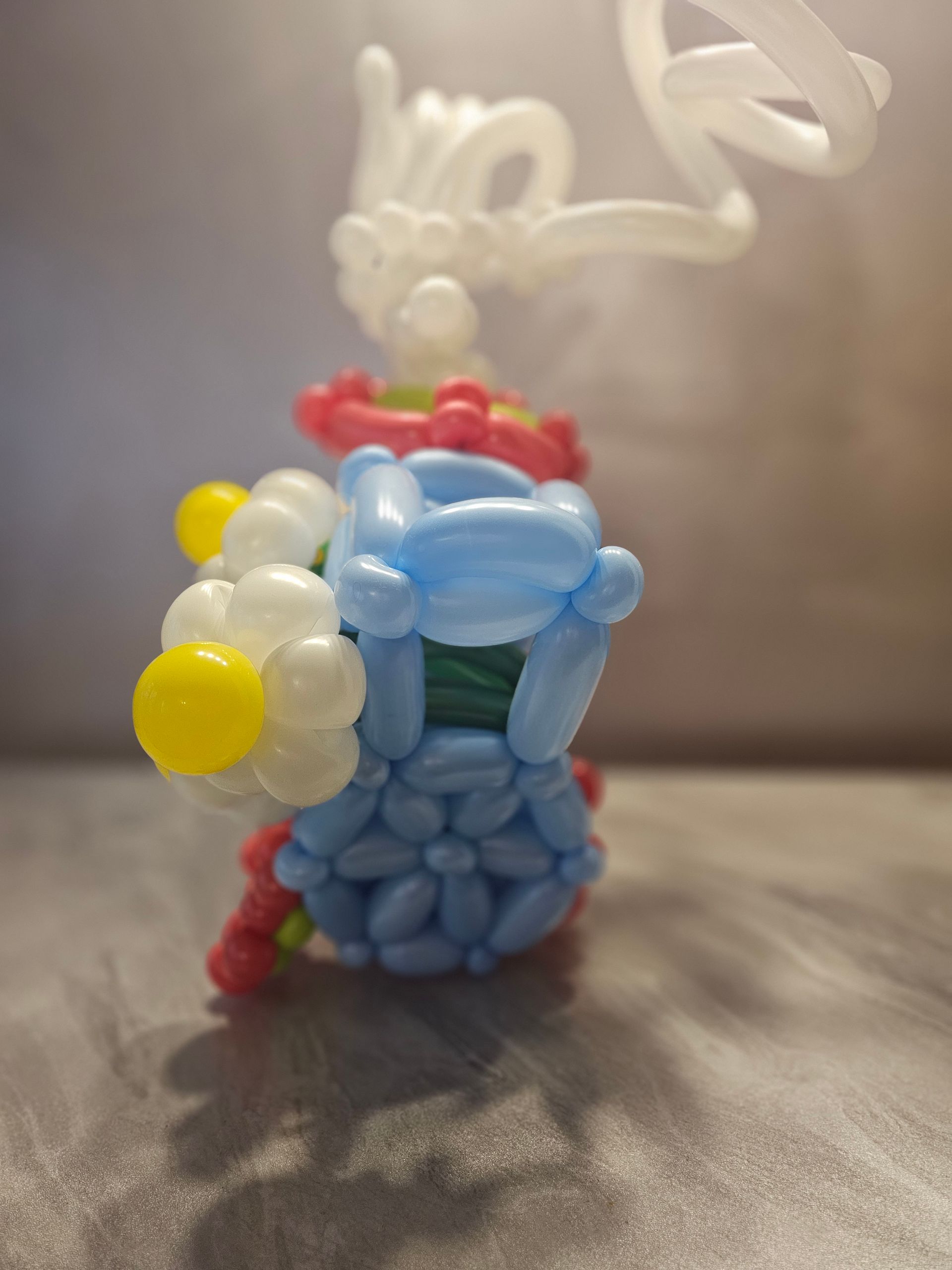 Balloon sculpture of a blue vase with flowers in pink, white, and yellow against a blurry background.