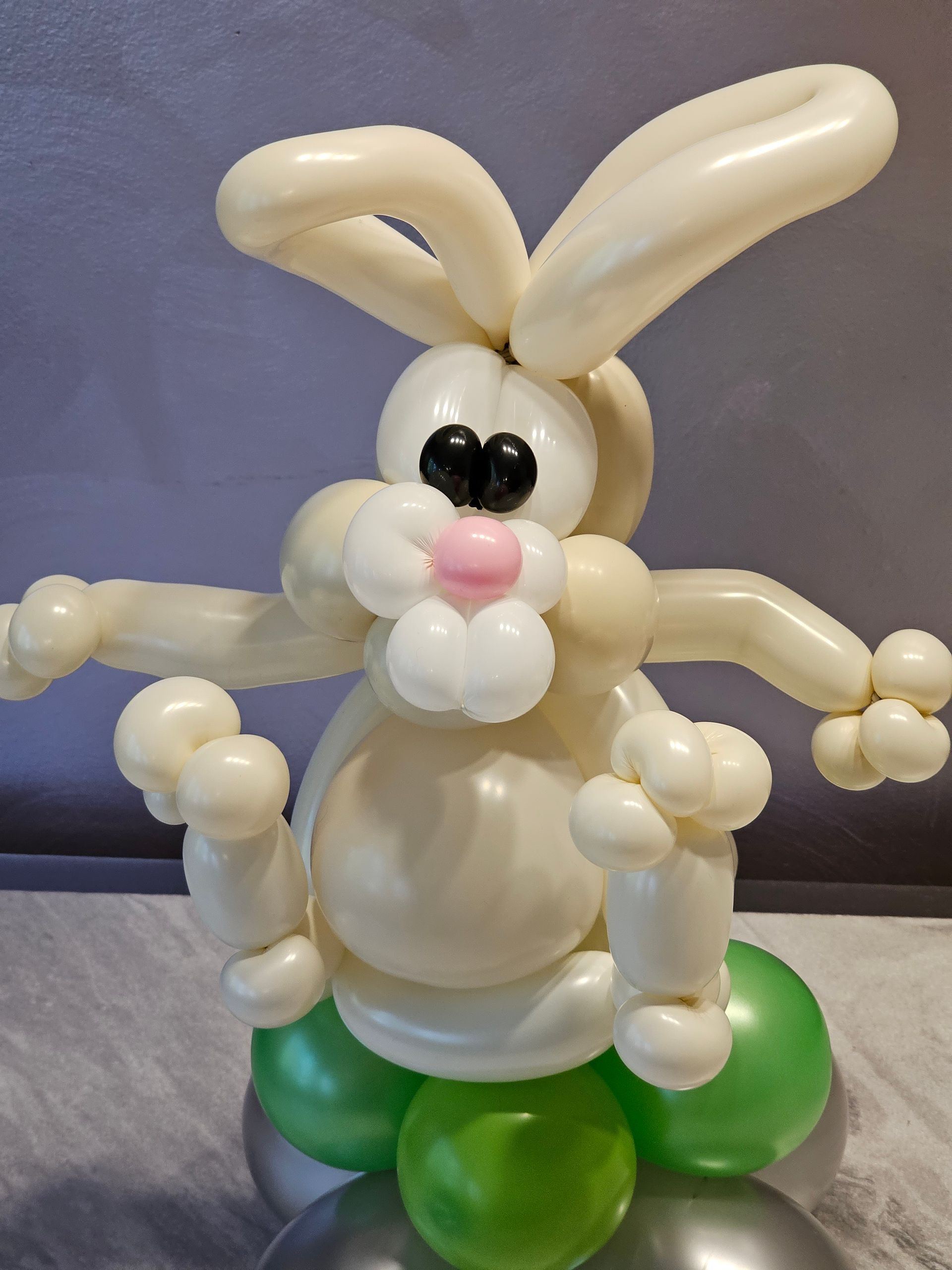 Balloon sculpture of a white bunny with outstretched arms atop green and silver balloons.