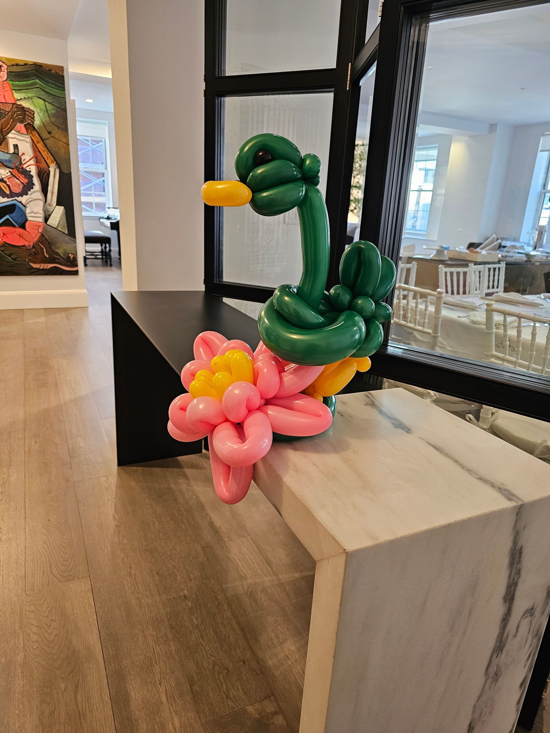Balloon sculpture of a green duck with a yellow beak perched on a pink and yellow flower; displayed on a marble bench.