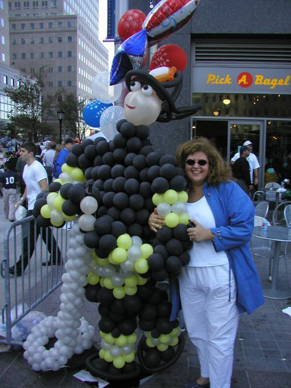 Woman with a balloon firefighter in front of Pick A Bagel; city setting, balloons overhead.
