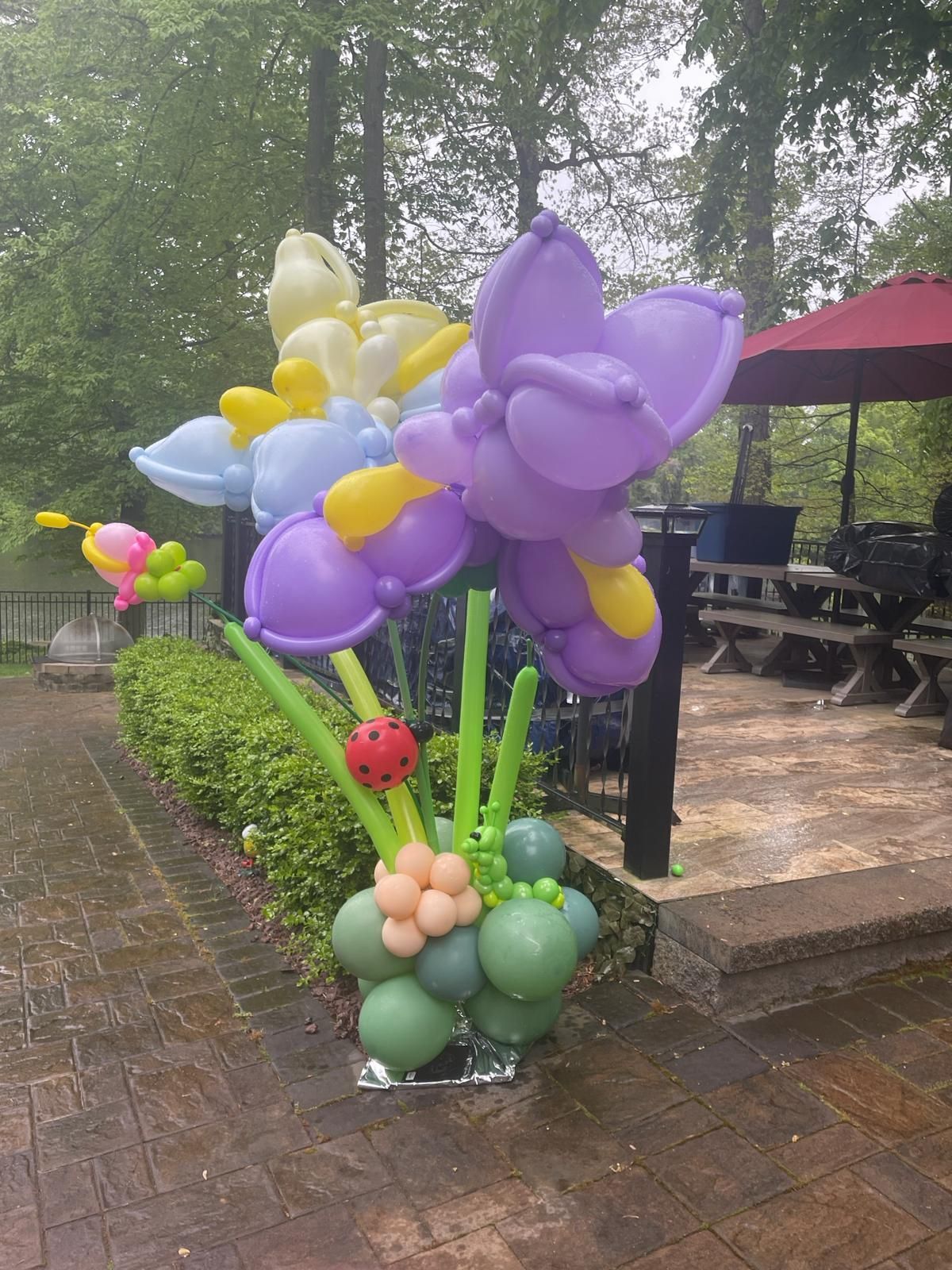 Balloon flower sculpture with purple, yellow, and blue petals, green stems, and a ladybug; set on a brick patio.