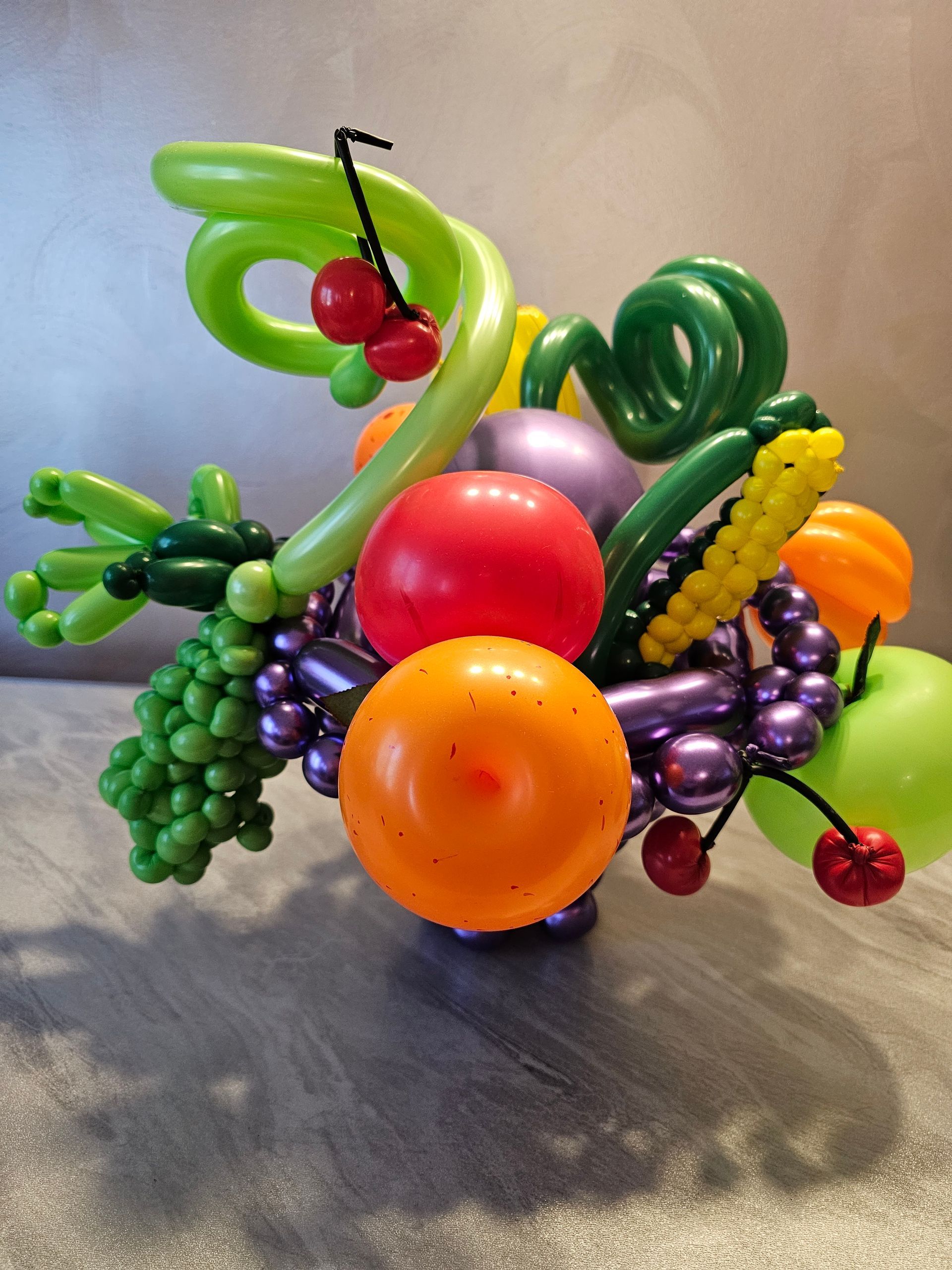 Balloon sculpture of a fruit basket; includes grapes, cherries, orange, apple, and other colorful fruits.
