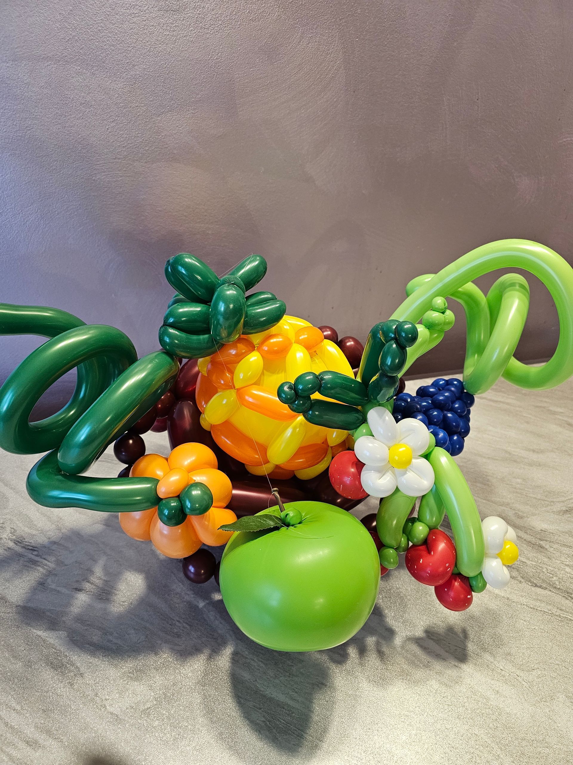Balloons crafted into a fruit basket with green vines, yellow pineapple, red berries, and green apple.