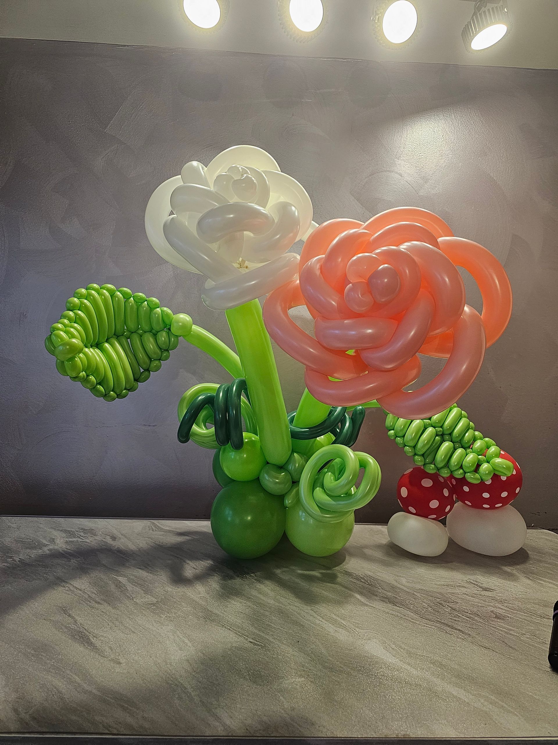 Balloon sculpture of flowers: white and pink roses, green stems and leaves, and a small mushroom.