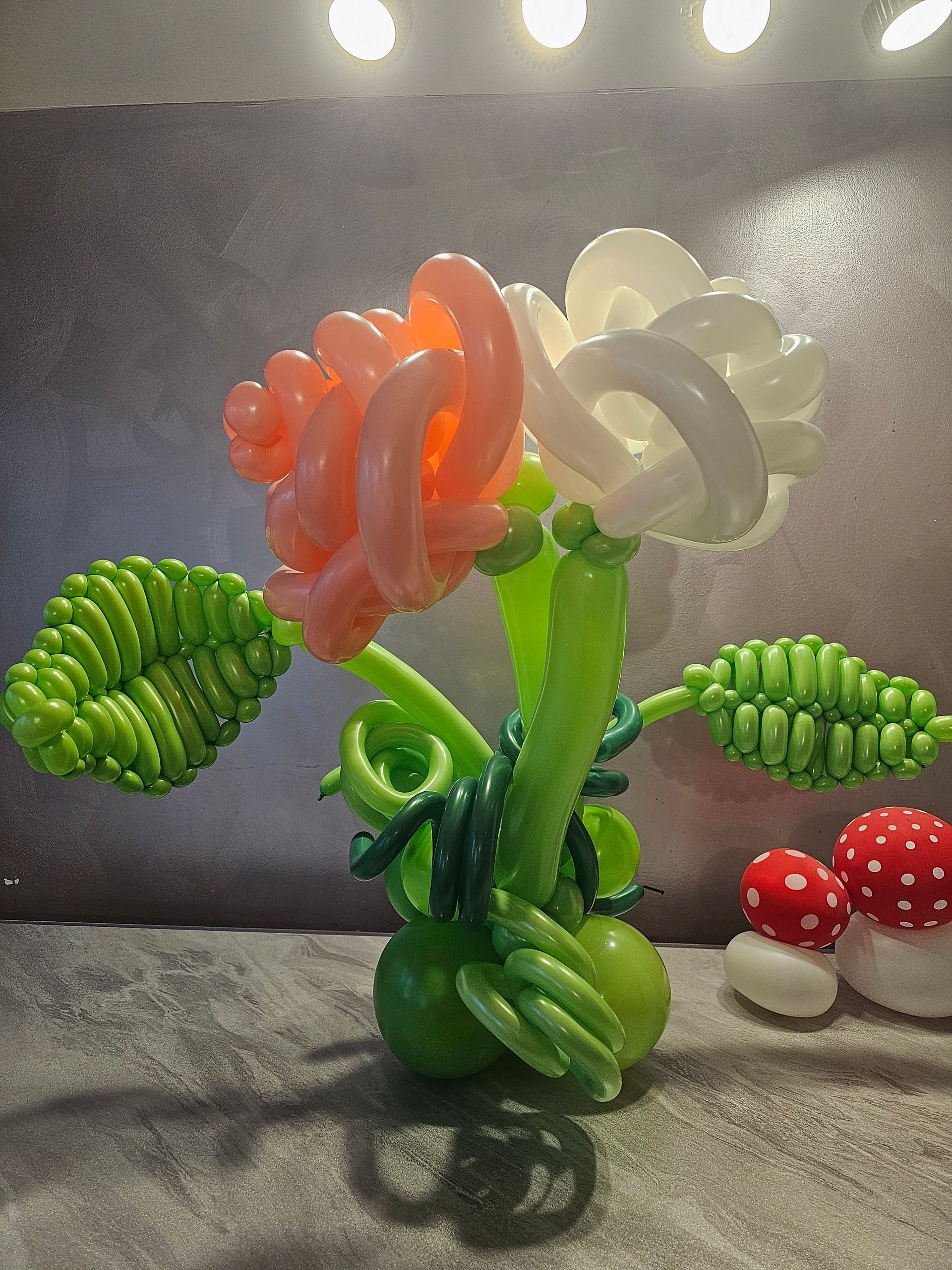 Balloon sculpture of a rose with orange and white petals, green stem and leaves, with small mushroom balloons.