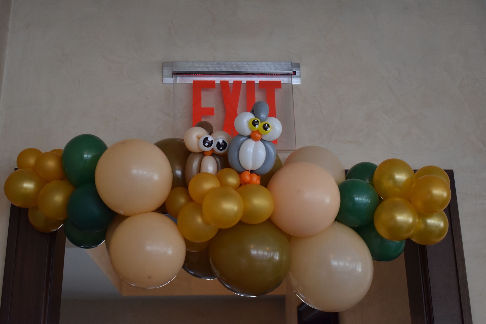 Balloon arch with two owl figures in front of an EXIT sign. Balloons are gold, green, and beige.