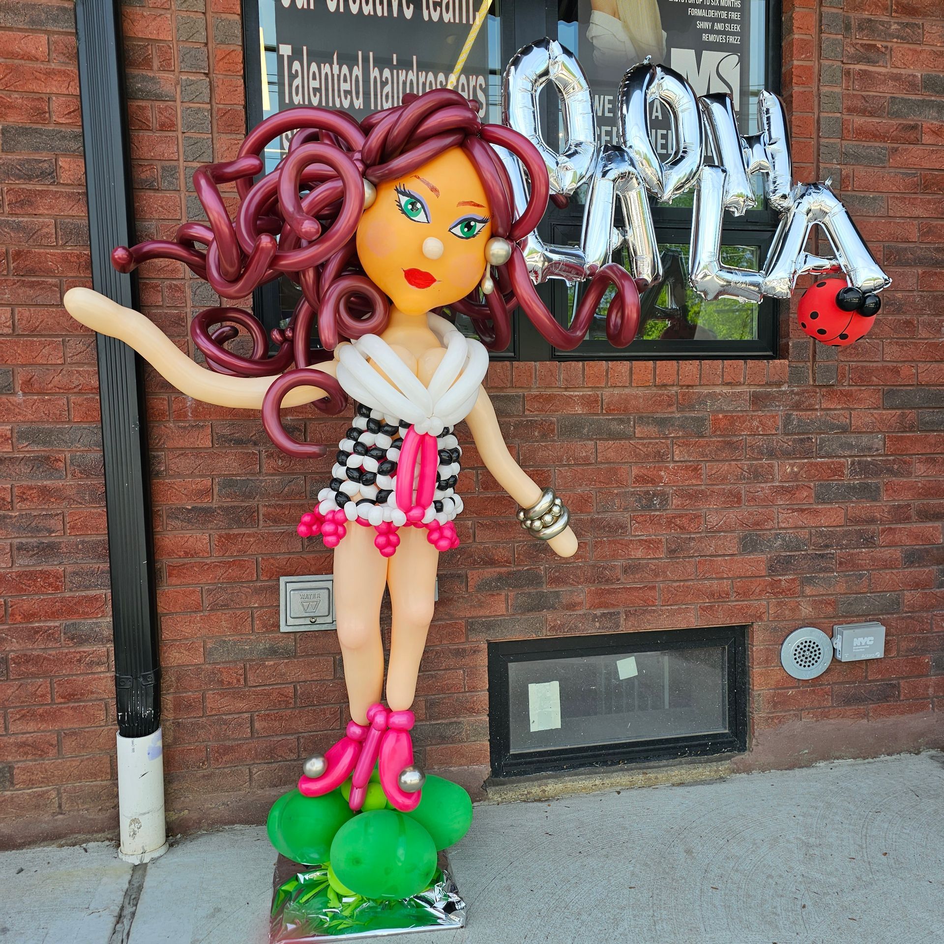 Balloon sculpture of a woman in front of a brick building. She is wearing a checkered dress and holding a ladybug.