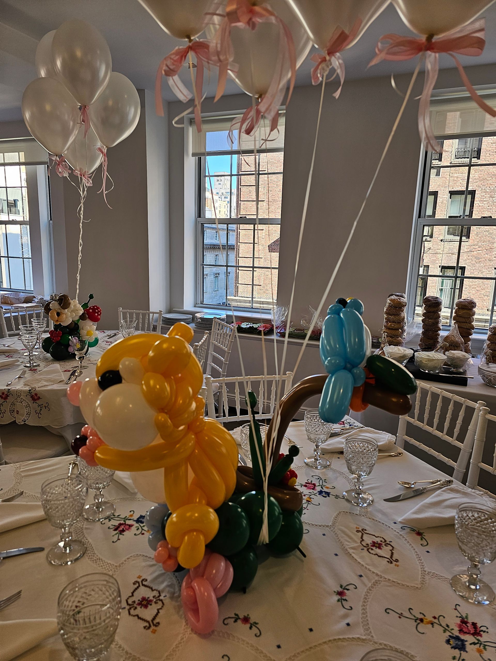 Table setting with balloon art: girl, bird, & bunches of white balloons with pink ribbons; windows in background.