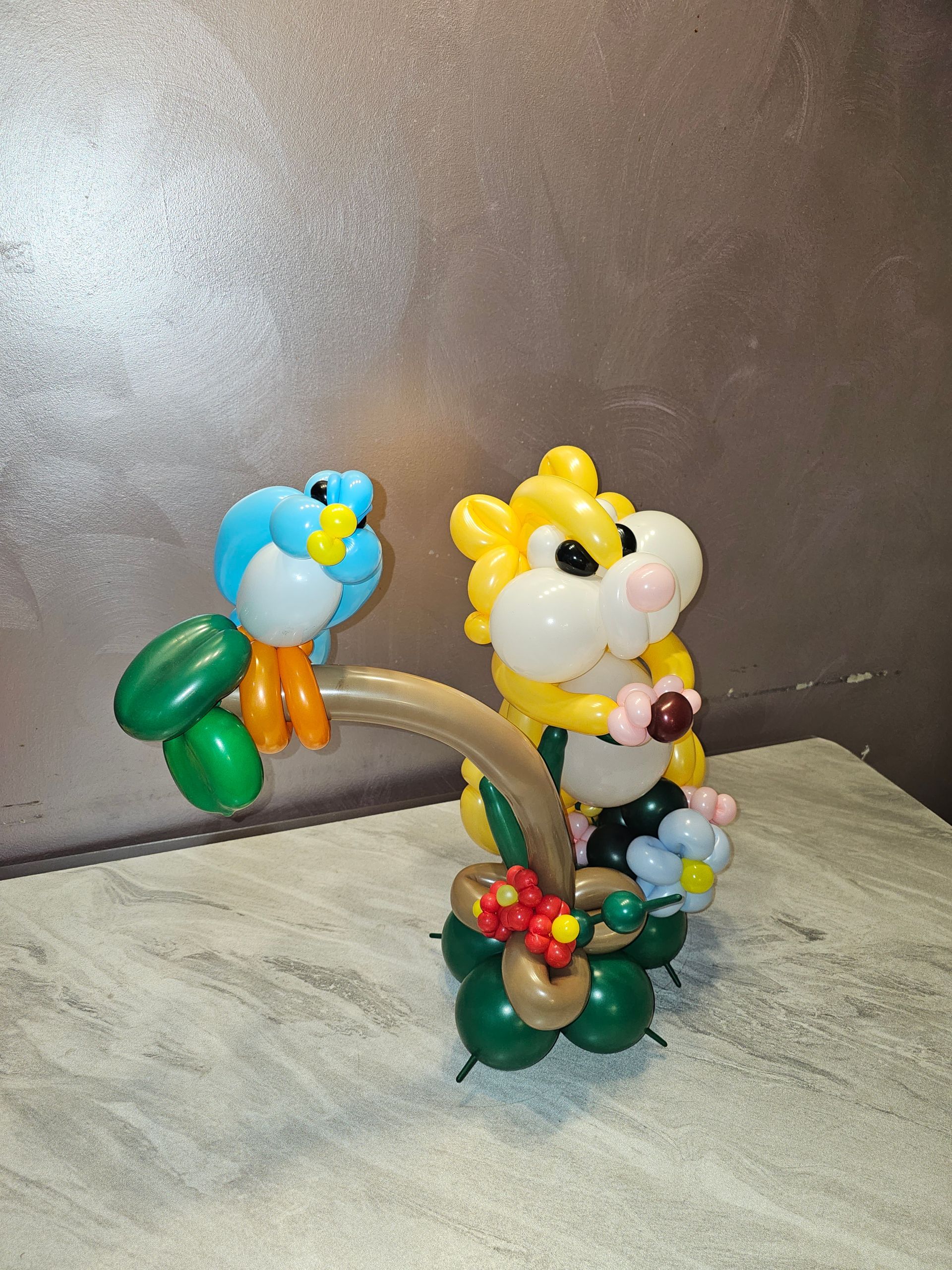 Balloon art of a blue bird perched on a brown vine next to a yellow hamster holding flowers.
