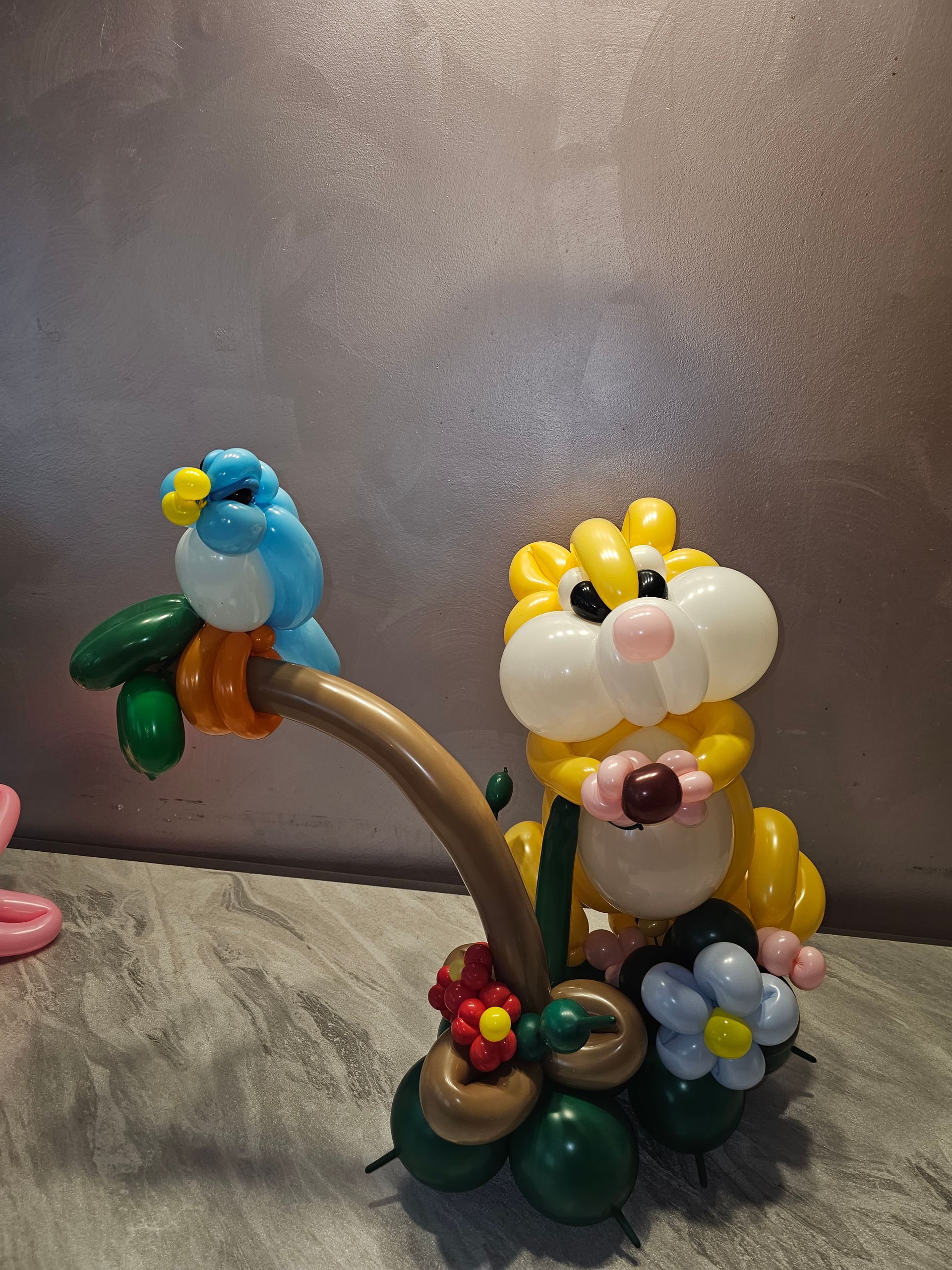 Balloon sculpture of a hamster with a blue bird on a branch, resting on a base with flowers and leaves.
