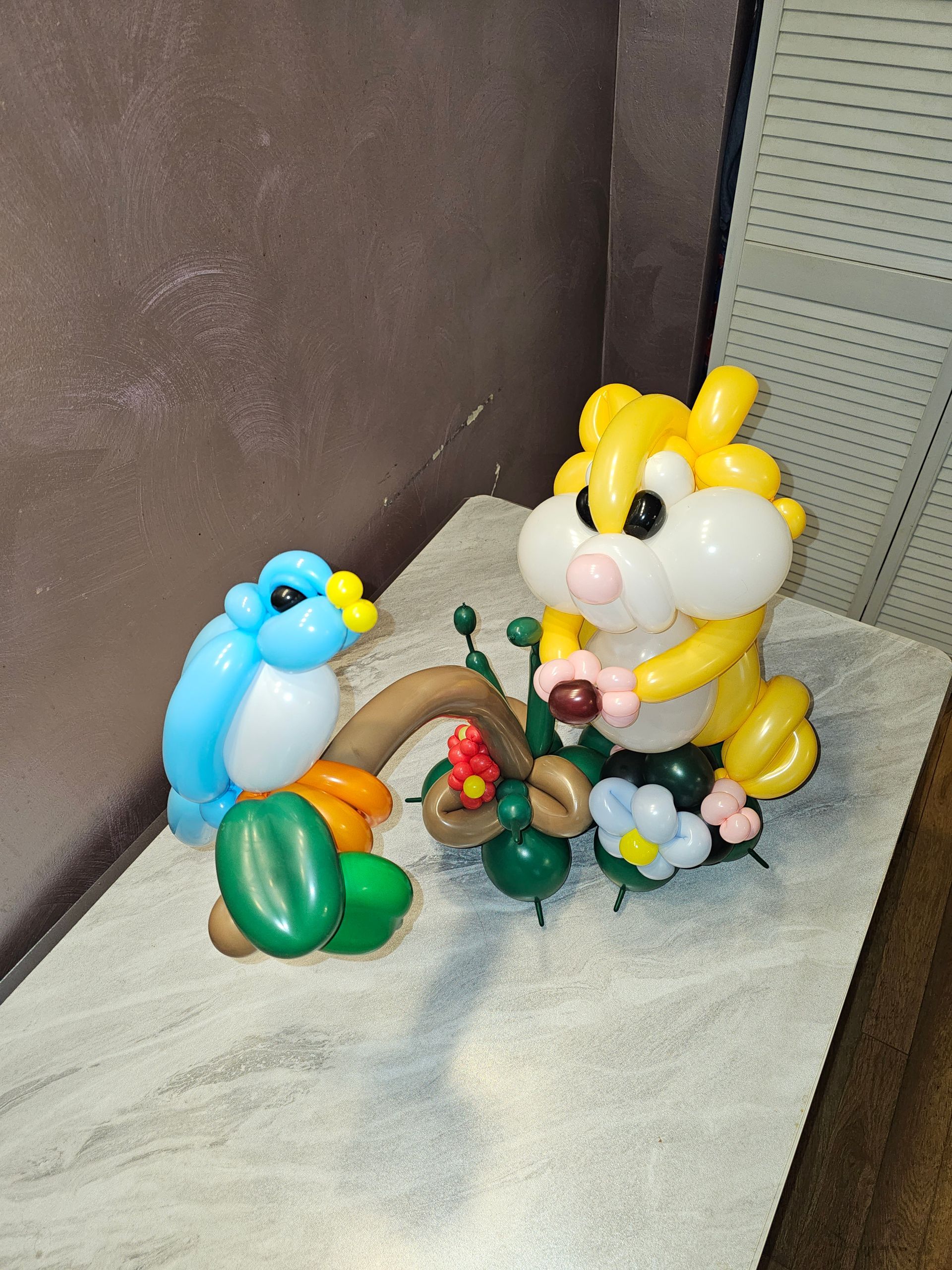 Balloon animal sculptures of a bluebird and a hamster, next to a cactus with flowers.