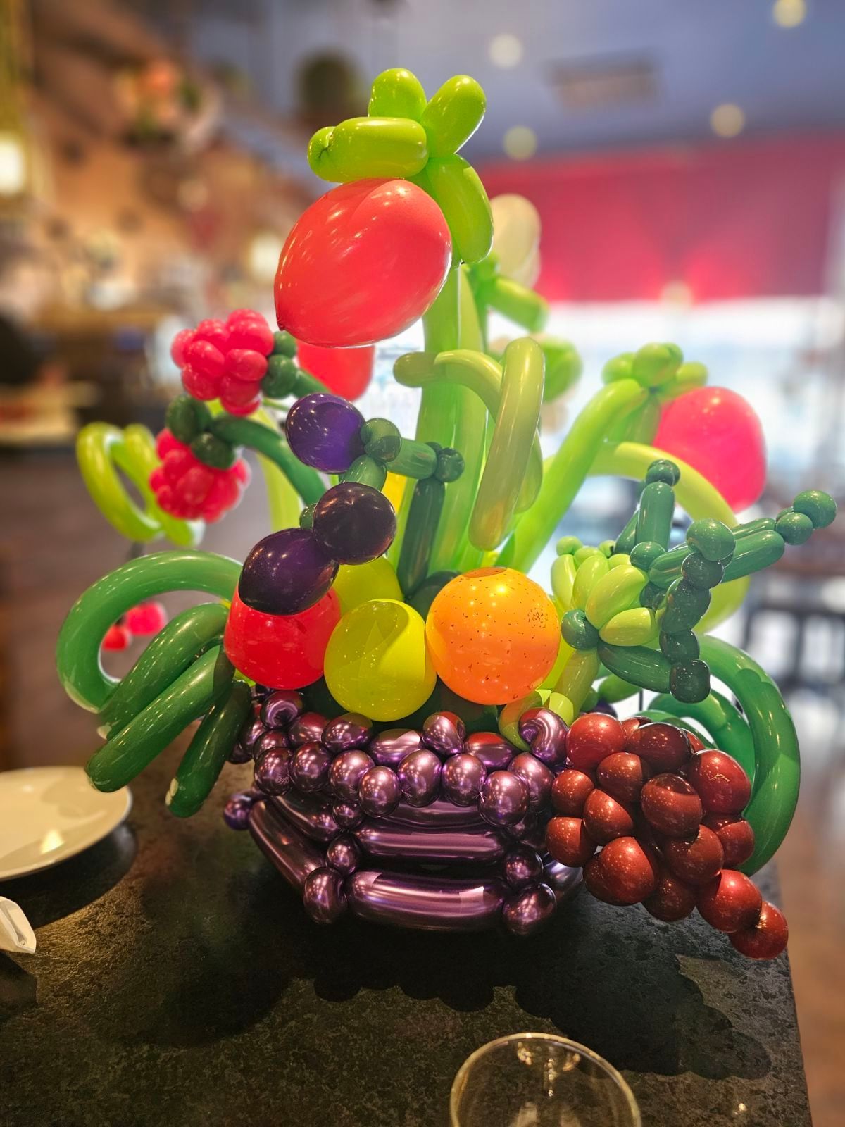 Balloon arrangement of colorful fruit; red, purple, orange, and green, in a purple basket.