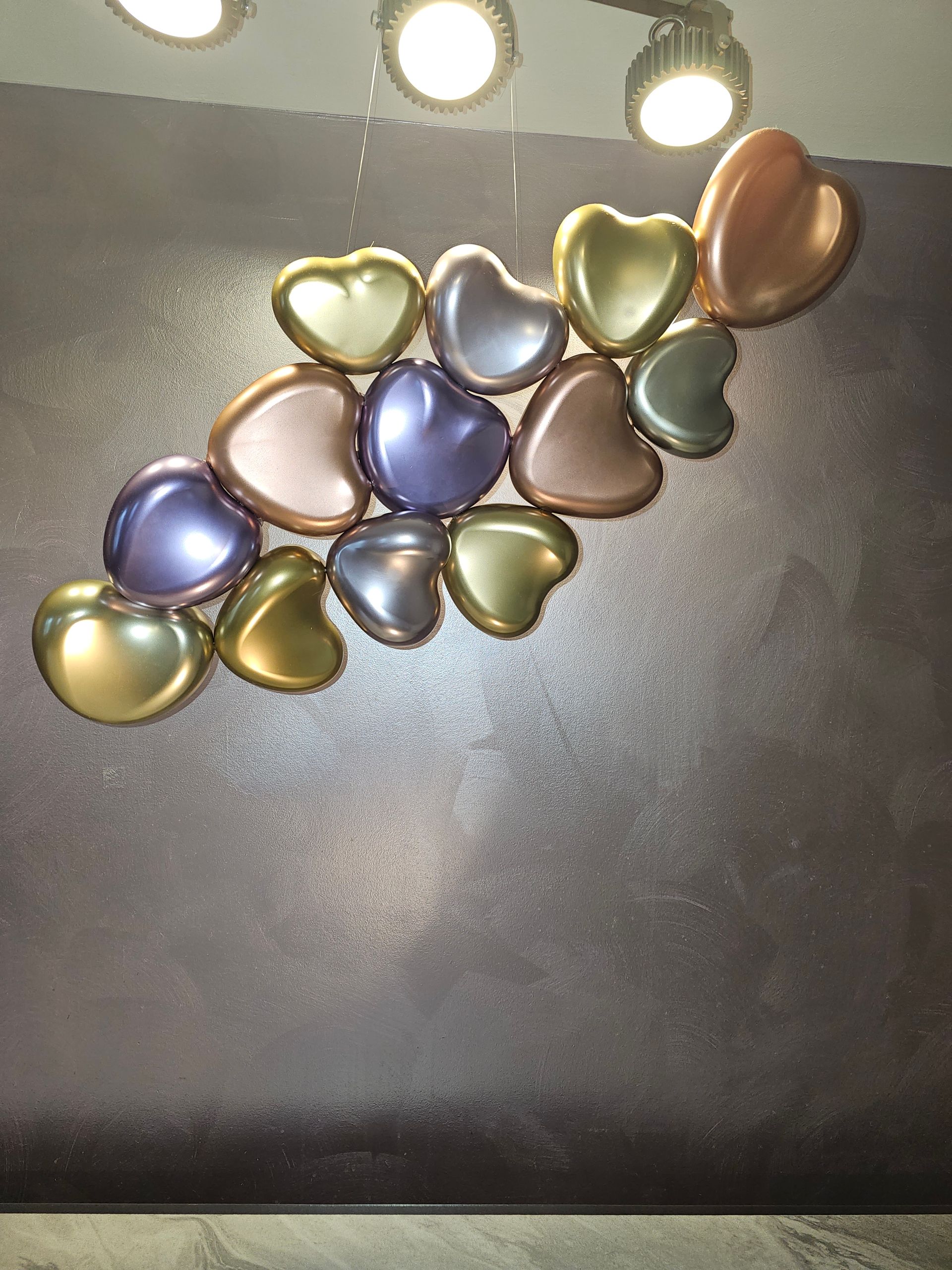Metallic heart-shaped wall art in various colors under three lights on a textured dark wall.