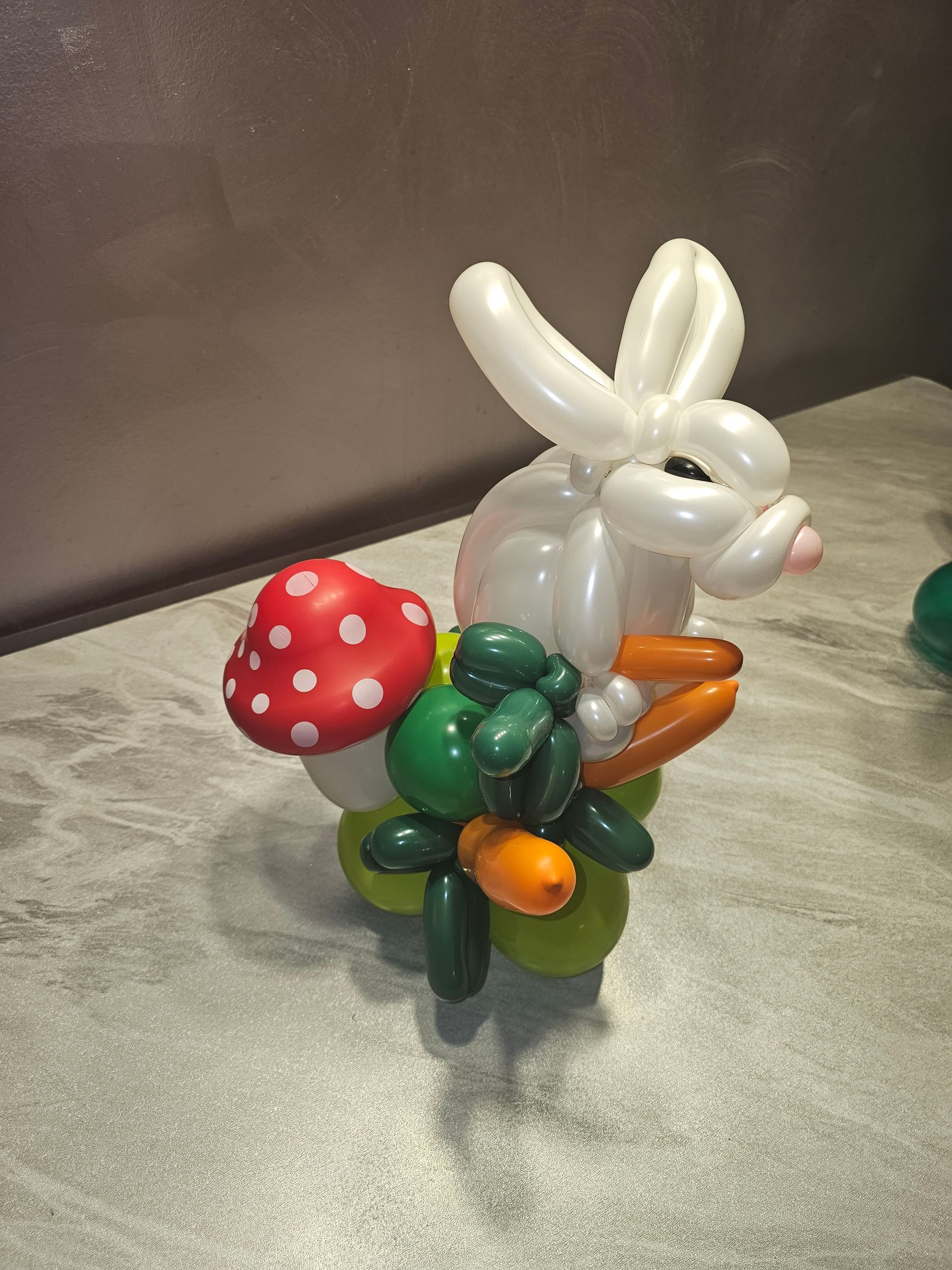Balloon sculpture of a white bunny holding carrots, with a mushroom and greenery on a green base.