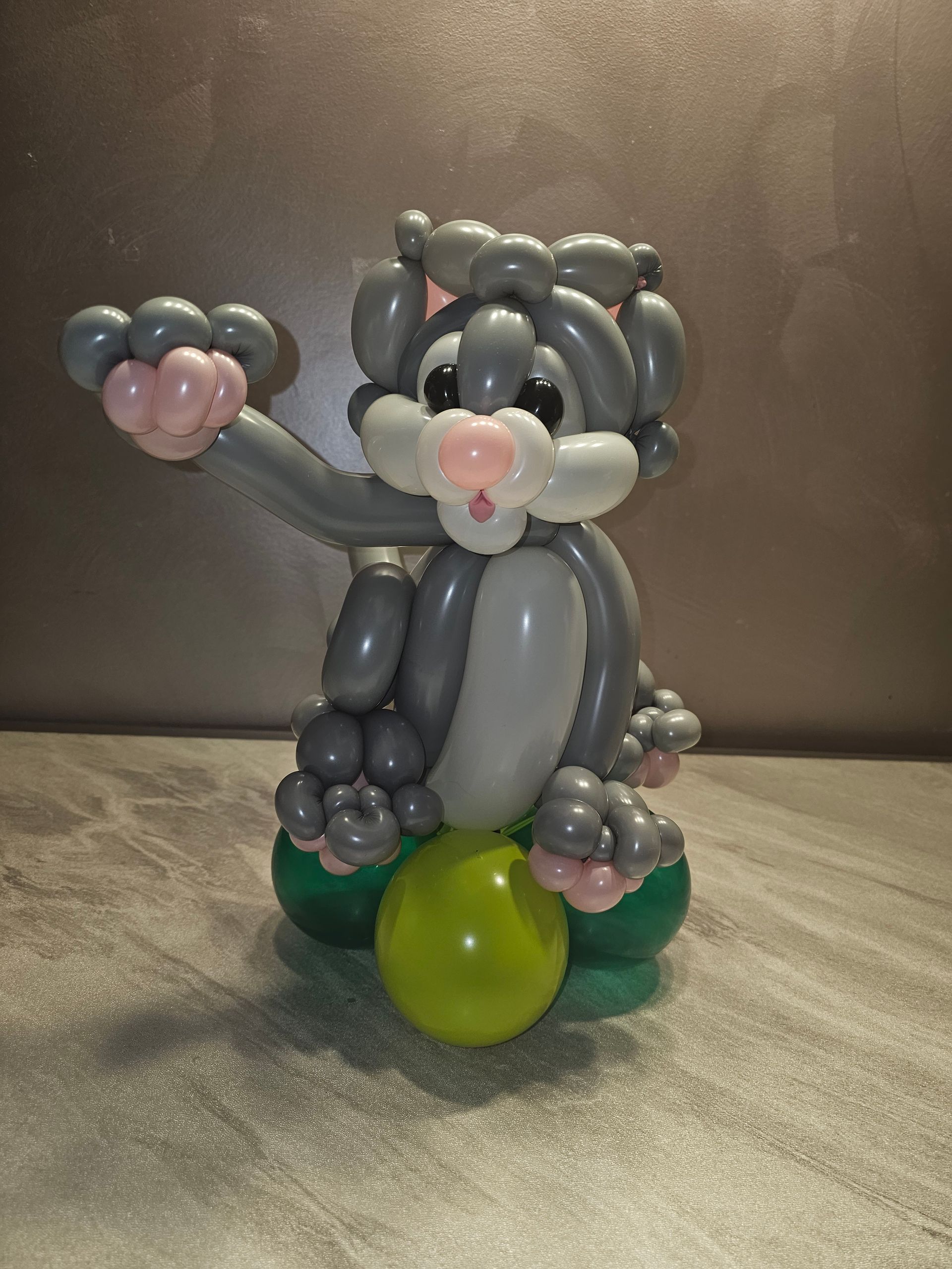 Balloon sculpture of a gray bunny with a pink nose, holding a green ball, on a tabletop.