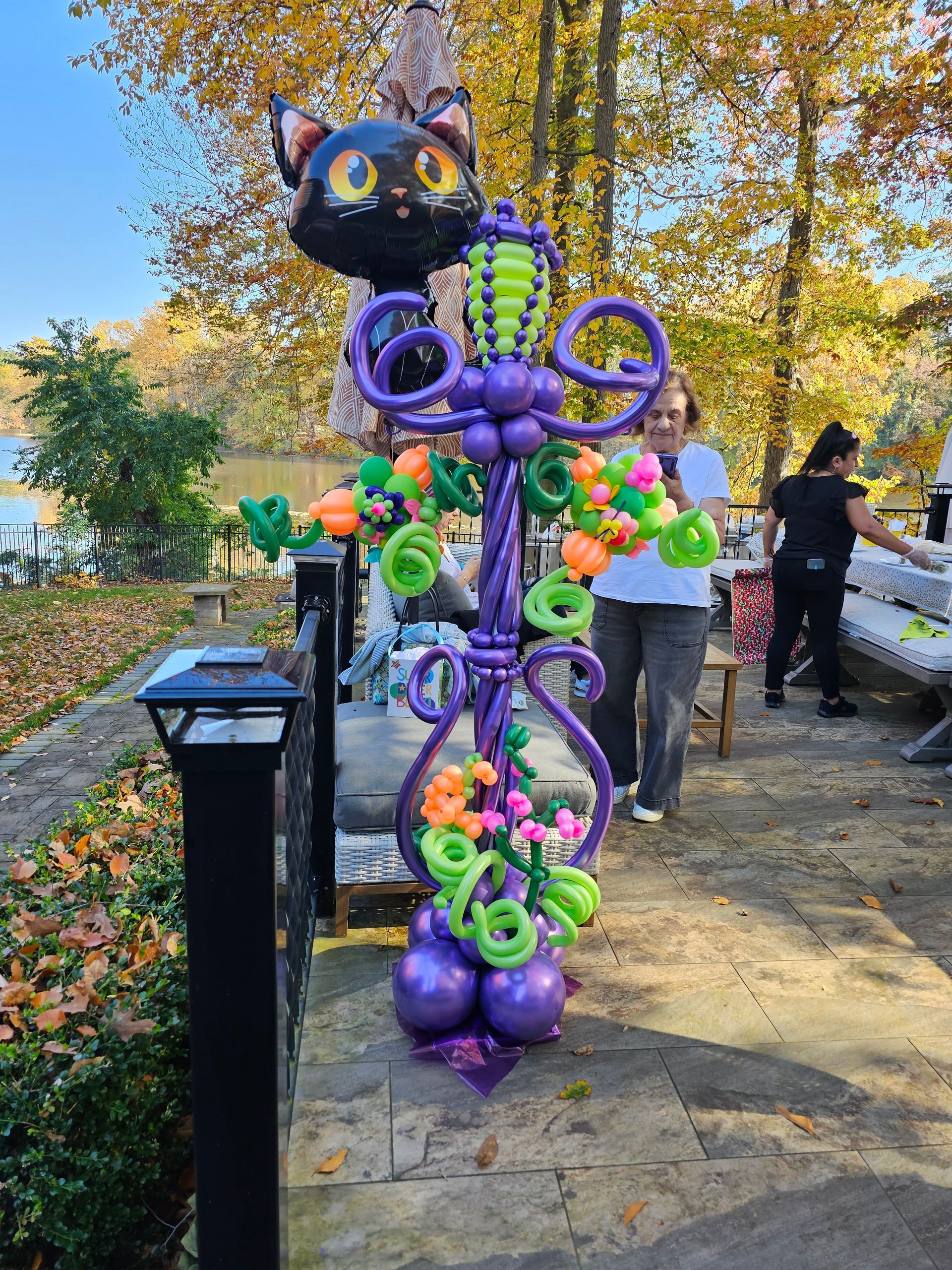 Halloween balloon sculpture of a black cat, purple base, flowers, and vines. Outdoors with people in the background.